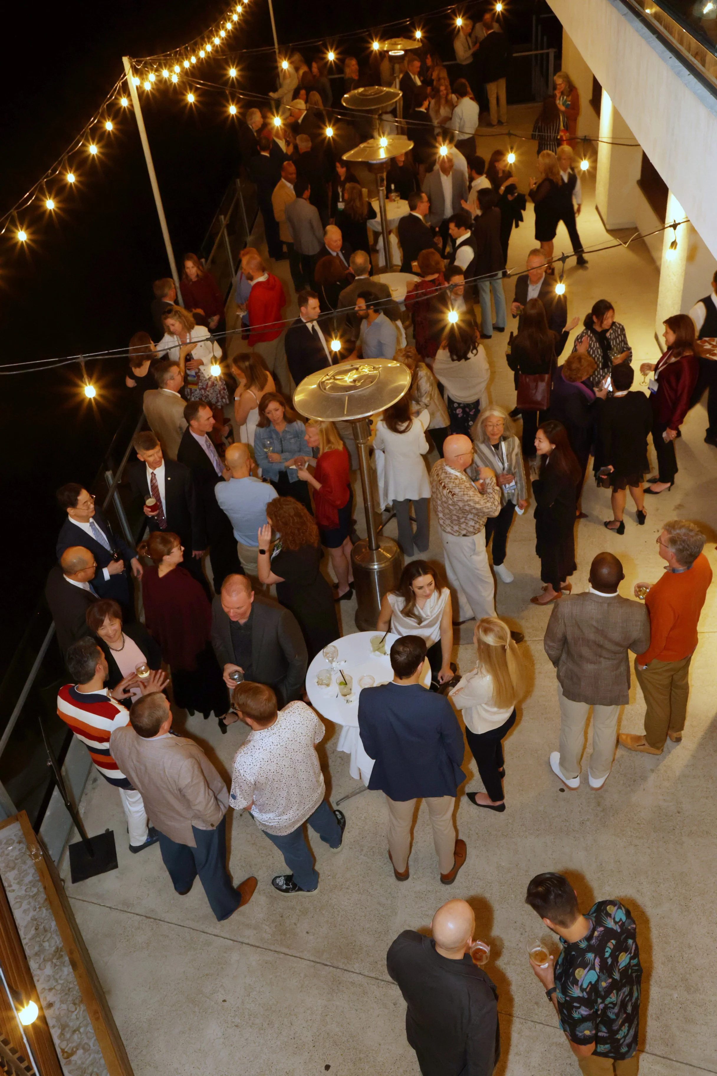 People socializing at an outdoor evening event with string lights overhead, some holding drinks, gathered around small tables and engaging in conversation.