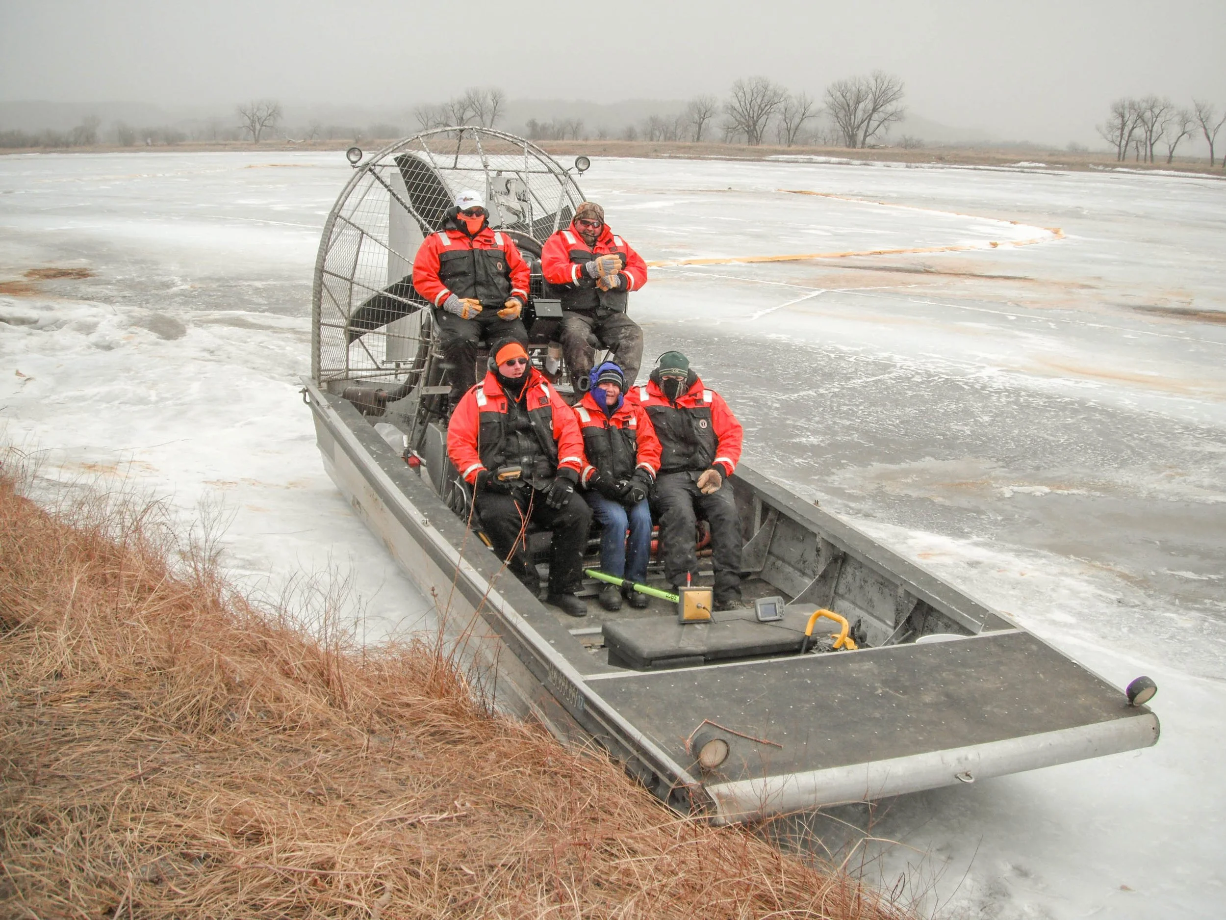 Five people wearing red jackets and winter gear riding a hovercraft over ice on a frozen body of water.