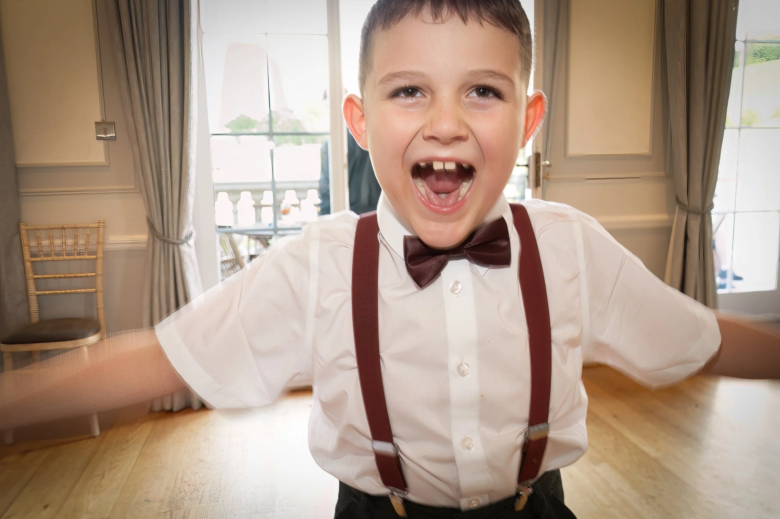 Young boy dressed in white shirt with suspenders and bow tie, smiling excitedly with arms outstretched in a well-lit room with large windows and cream curtains.
