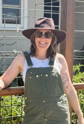 woman in green overalls and white tank top with brown hat standing in front of garden