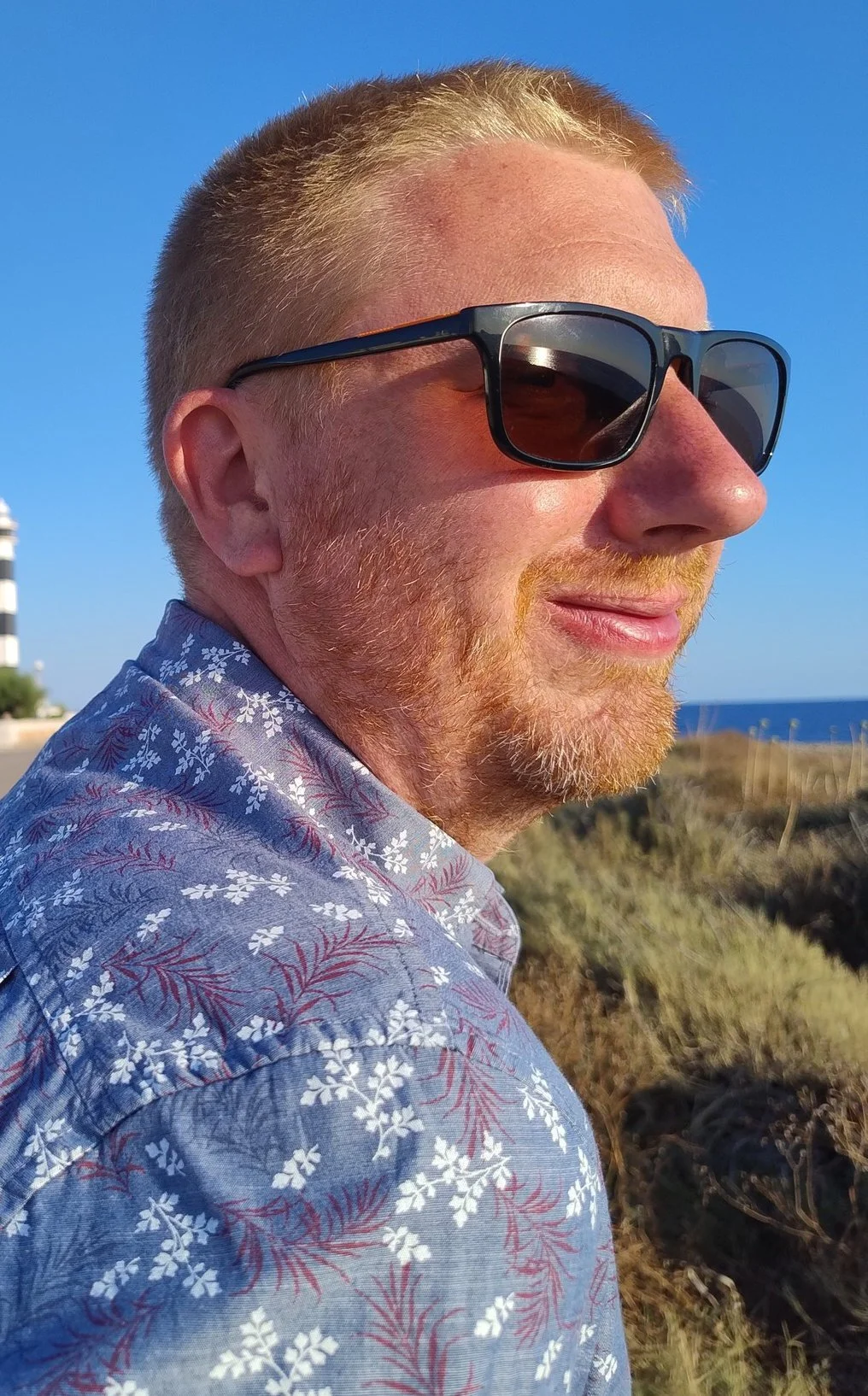 Ian has short blonde hair and has sunglasses on, behind is a blue sunny sky
