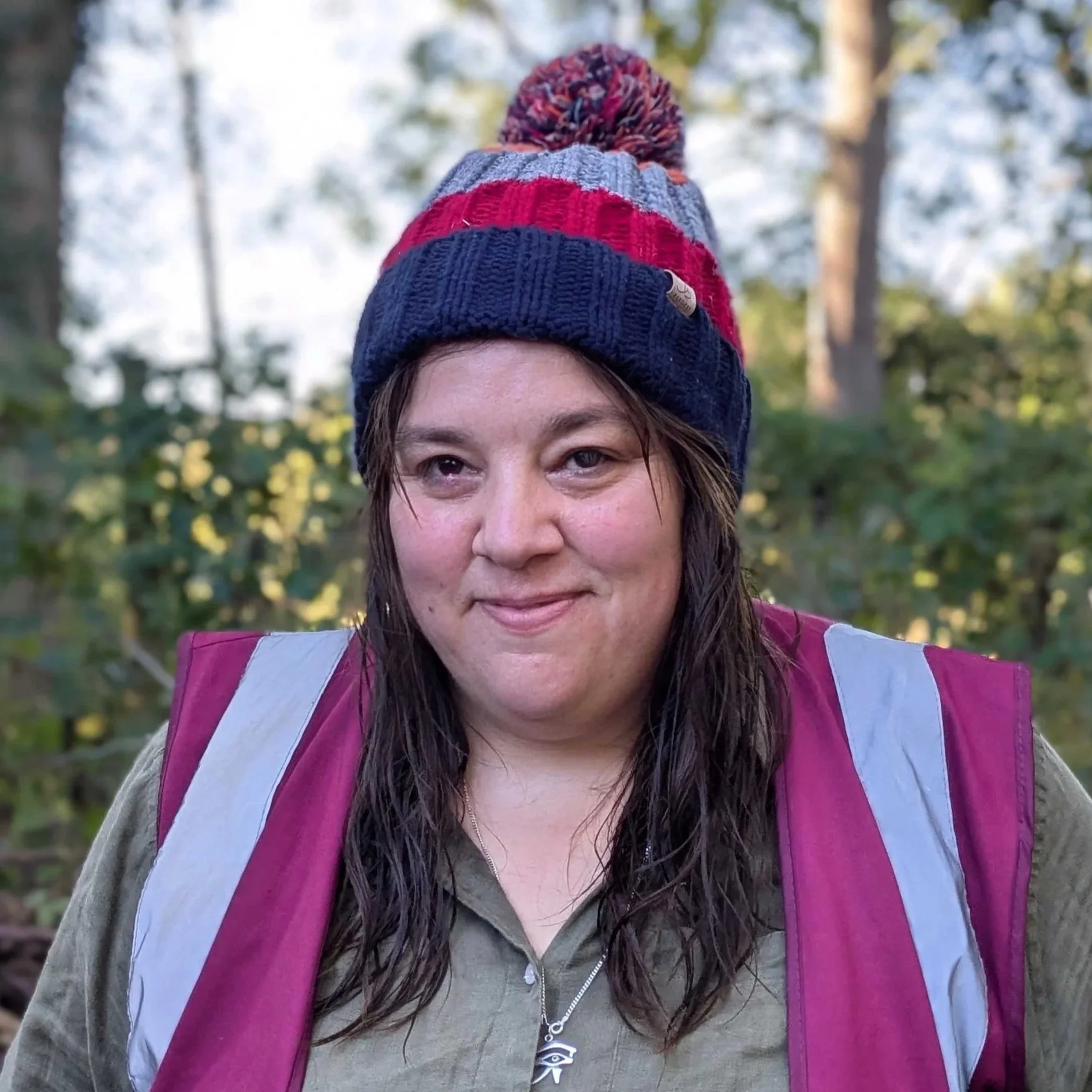 A woman smiling outdoors, wearing a black and pink North Face jacket and a blue shirt, with a pink backpack, short brown hair, and trees in the background.