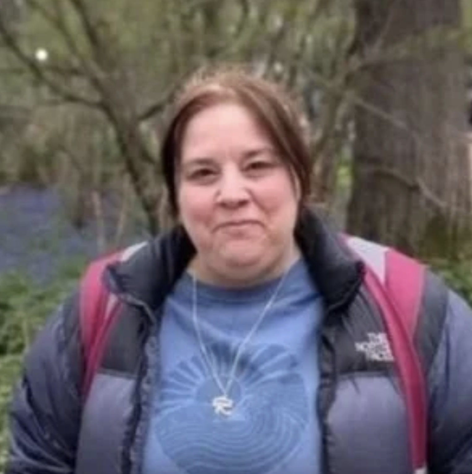 A woman smiling outdoors, wearing a black and pink North Face jacket and a blue shirt, with a pink backpack, short brown hair, and trees in the background.