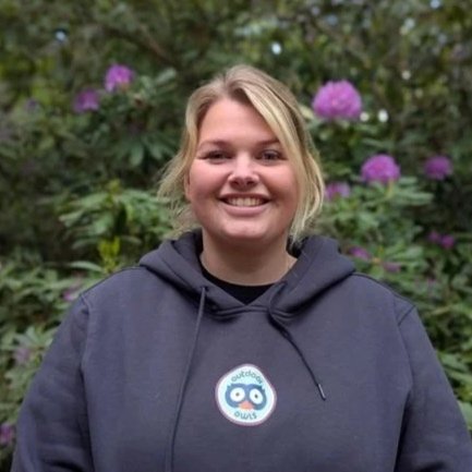 Young woman smiling outdoors with purple flowers and greenery in the background, wearing a dark hoodie with a logo.