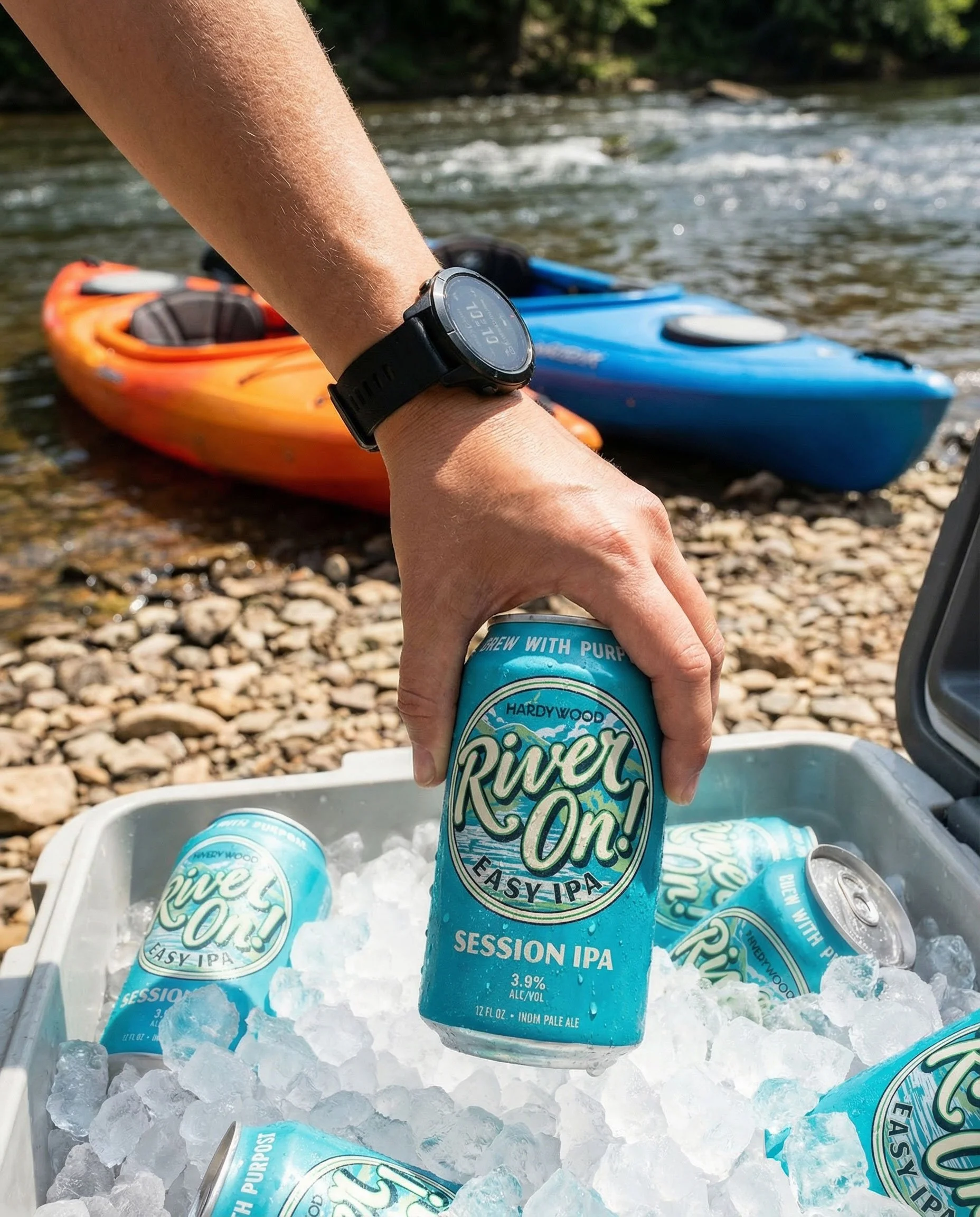A hand pulling an ice cold River On can out of a cooler filled with ice and River On cans next to a river bank with two kayaks.