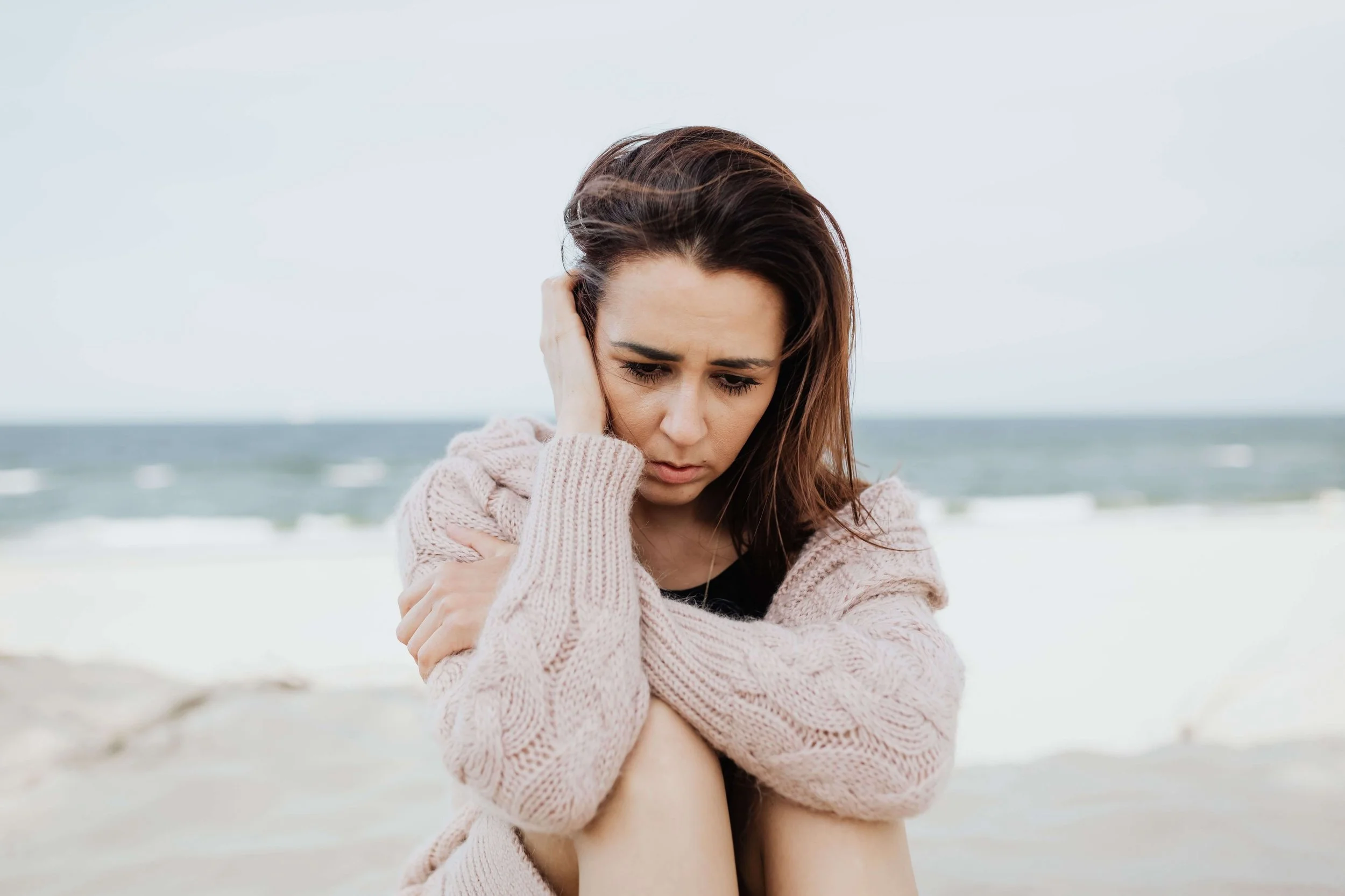 Woman sitting on the beach looking sad, representing the emotional struggles that can arise during the summer and the support available through therapy in Bay Shore, NY.