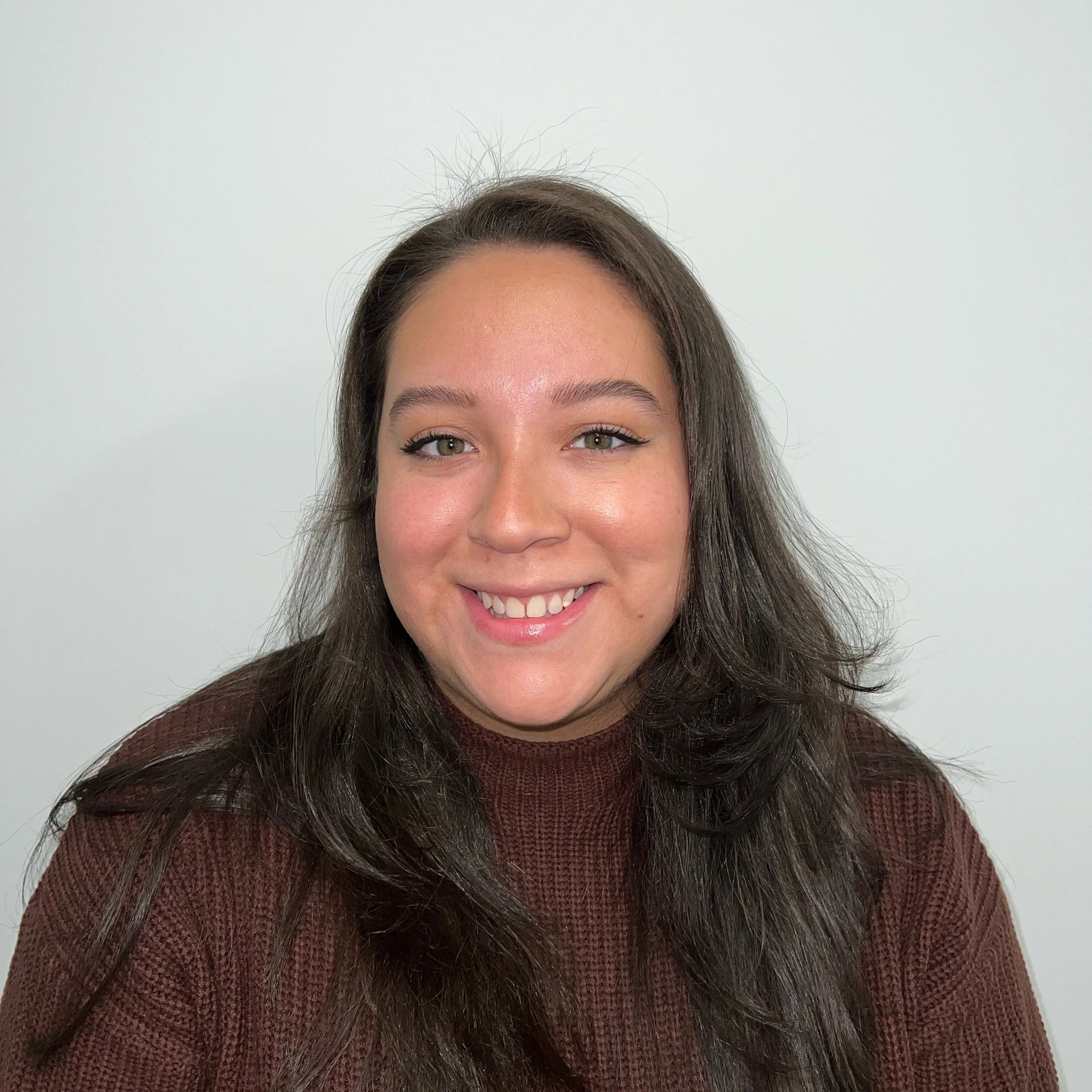 Melissa Reyes Martinez, a therapist, smiling warmly while wearing a brown sweater in a calm, welcoming setting. Trauma counseling for young adults in West Babylon, NY, can help if you’re struggling.