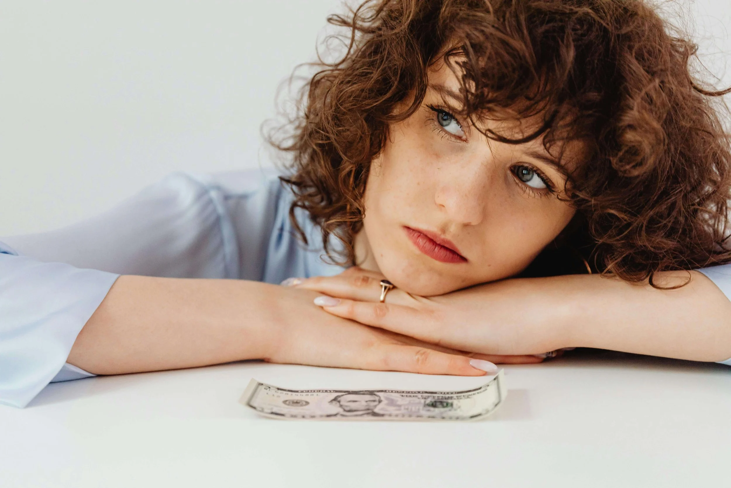 Young woman resting her head on a table with a five-dollar bill in front of her, representing financial pressure addressed in stress therapy for young adults in Bay Shore, NY.