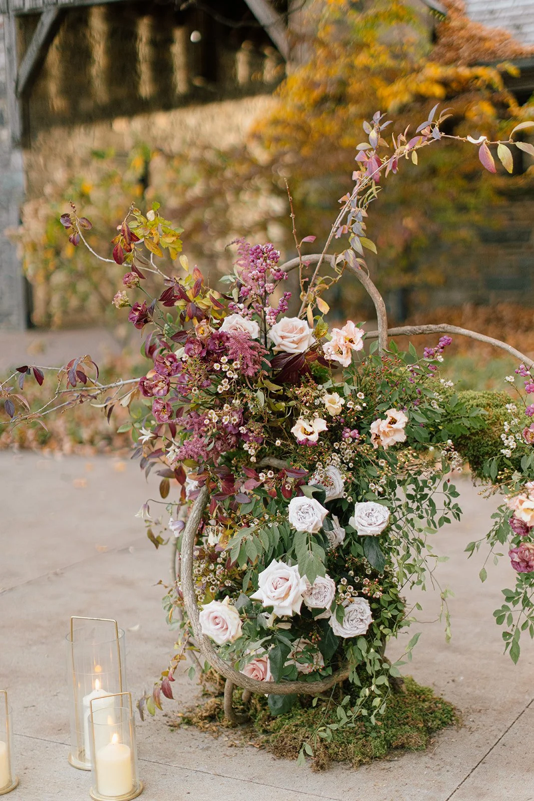 Autumn-themed floral arrangement with roses and colorful leaves, accompanied by lit candles in glass holders.