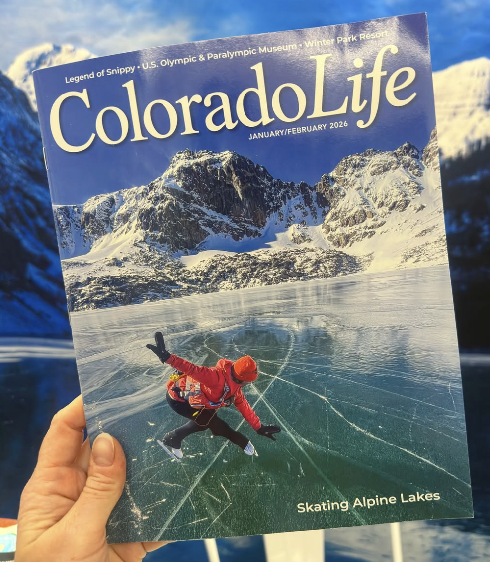 A person holding a Colorado Life magazine with a cover photo of an ice skater on a frozen lake with snow-covered mountains in the background, titled 'Skating Alpine Lakes' for January/February 2026.
