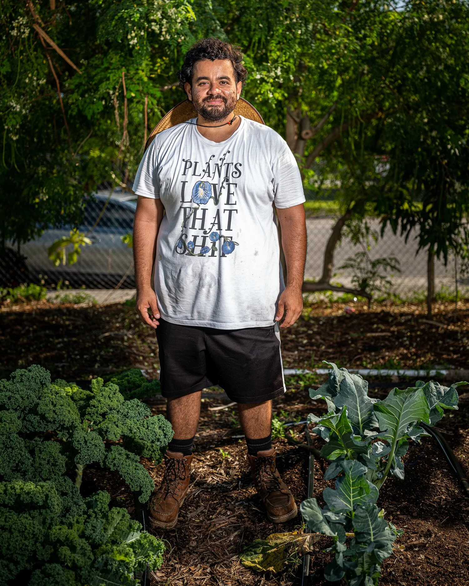 A man with curly hair, beard, and wearing a white T-shirt with floral text stands in a garden with leafy green vegetables. He's smiling, wearing brown boots, black shorts, and a conical straw hat resting on his back.