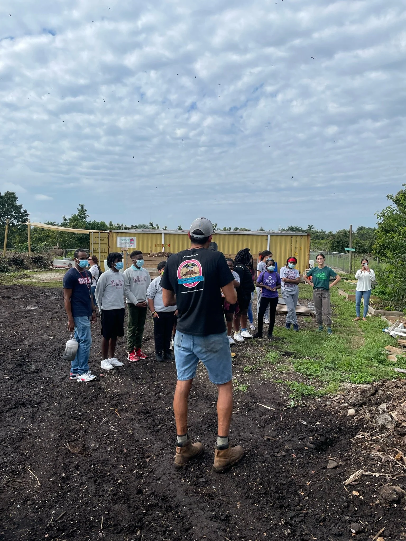 Group of people gathered outdoors on a cloudy day, listening to a person in a black T-shirt giving a presentation, with some wearing masks, in a garden or community space.