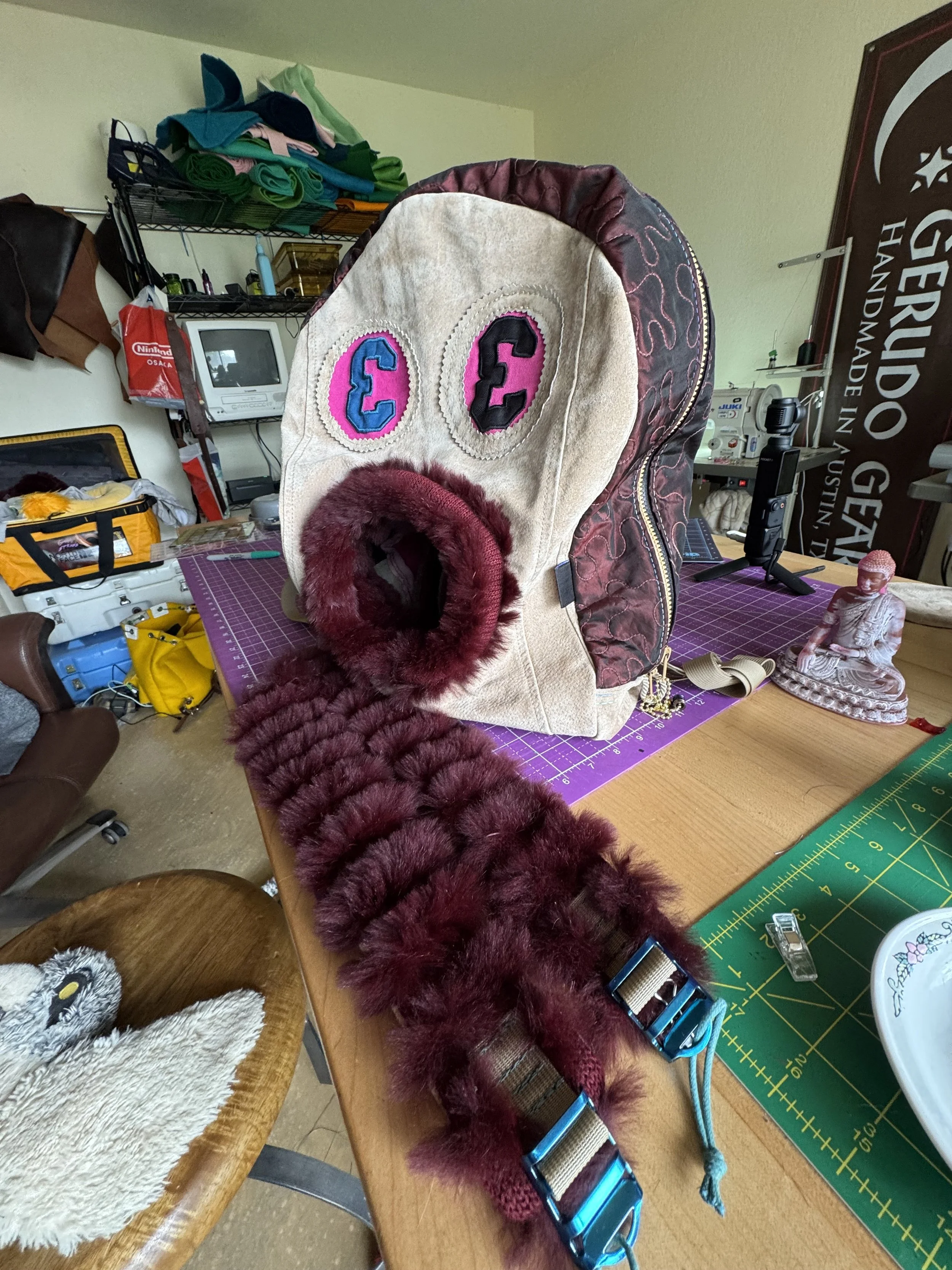A handmade hat resembling a face with embroidered eyes and a furry scarf, placed on a cluttered worktable in a craft room.
