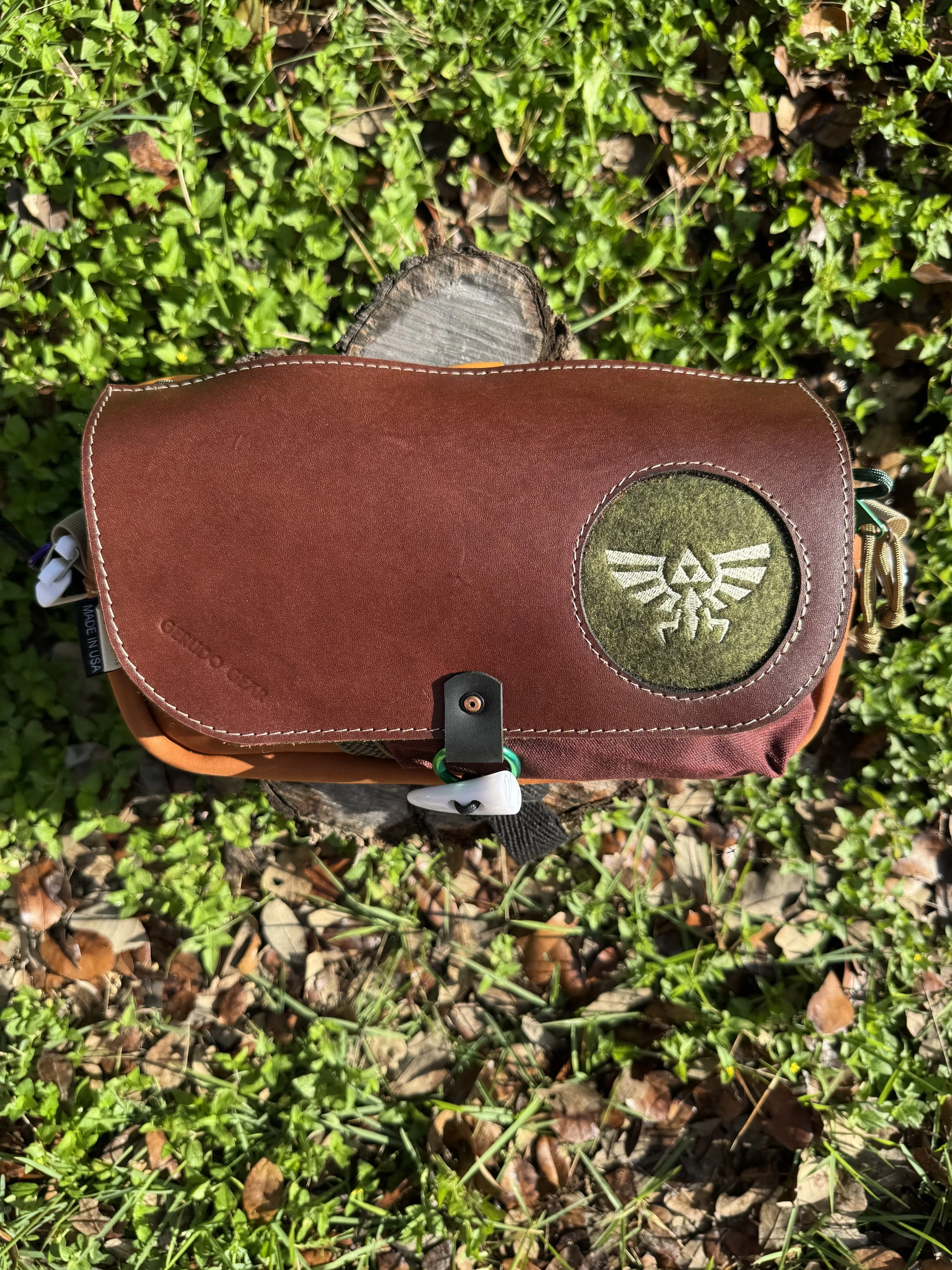 A brown and green Zelda-themed bag resting on a tree stump amongst green grass and dried leaves.