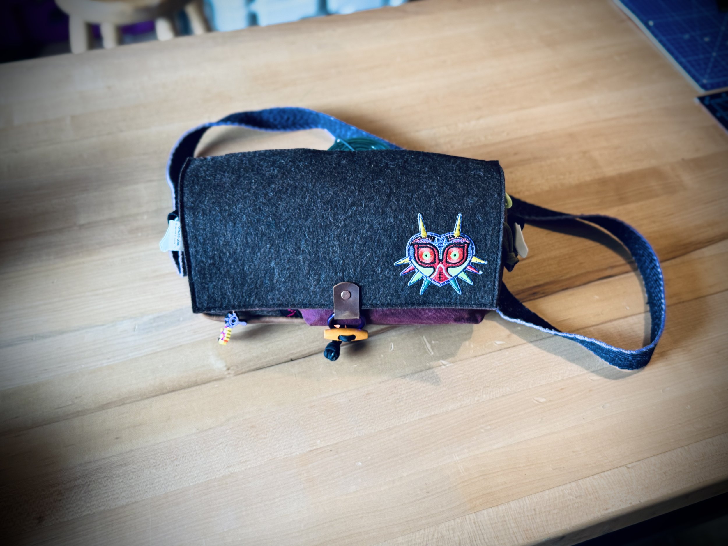 A small, handheld bag with a felt-like flap featuring a colorful embroidered mask design, resting on a wooden surface.