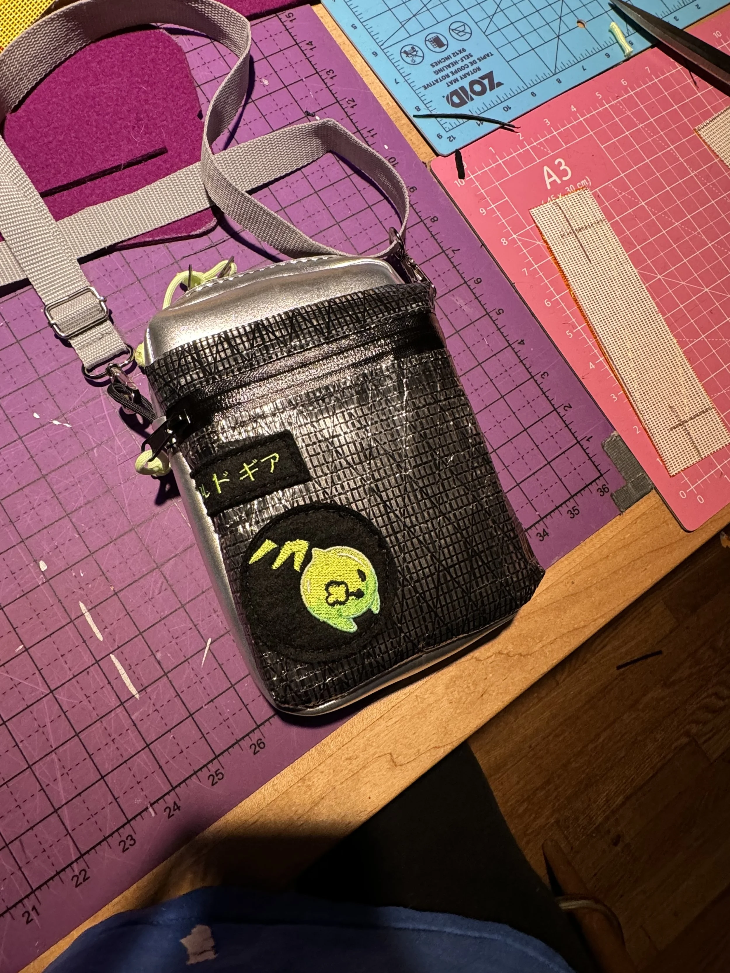 A small, black, and silver crossbody bag with yellow embroidered patch of a character with a speech bubble and Japanese characters on a purple cutting mat with various rulers and cutting tools.