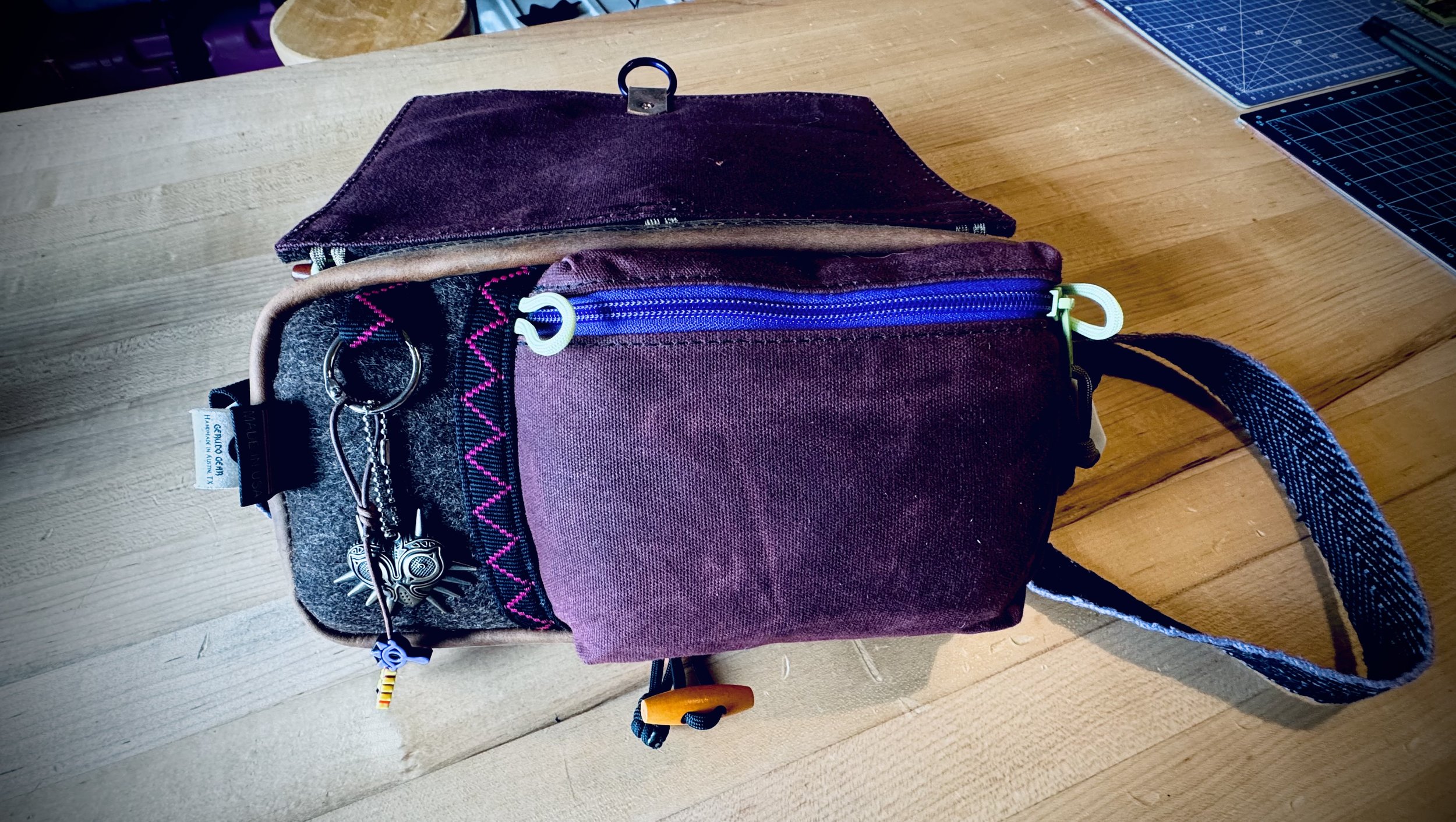 A small shoulder bag with purple and gray fabric, featuring a blue zipper on the front, placed on a wooden surface. The bag has keychains attached, including a metal night owl and a small rainbow-colored object. There is a fabric loop on the side and