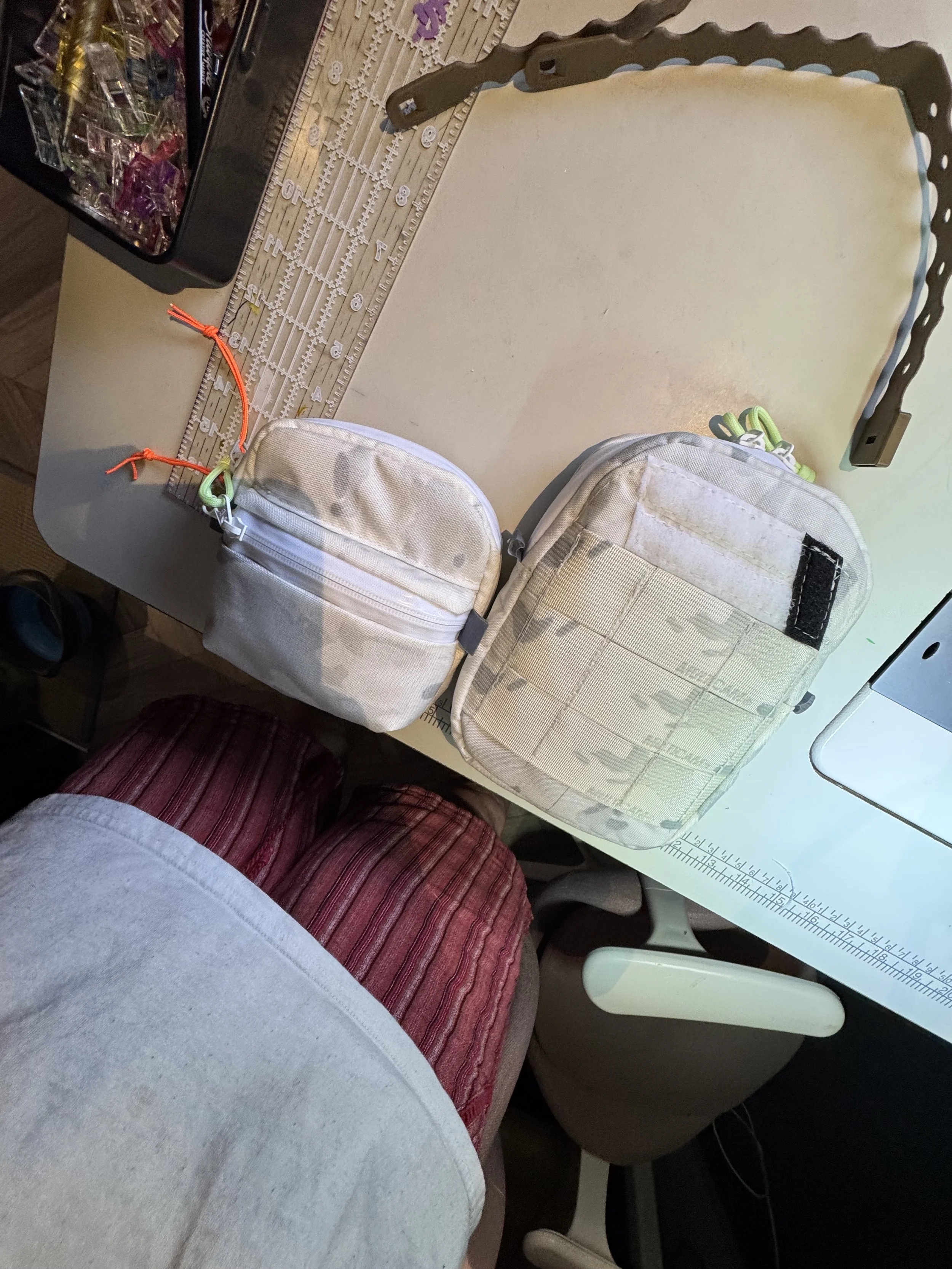 Two beige tactical backpacks on a white desk, with a ruler, a container of small craft supplies, and a person's legs in red striped shorts and a light gray shirt visible.