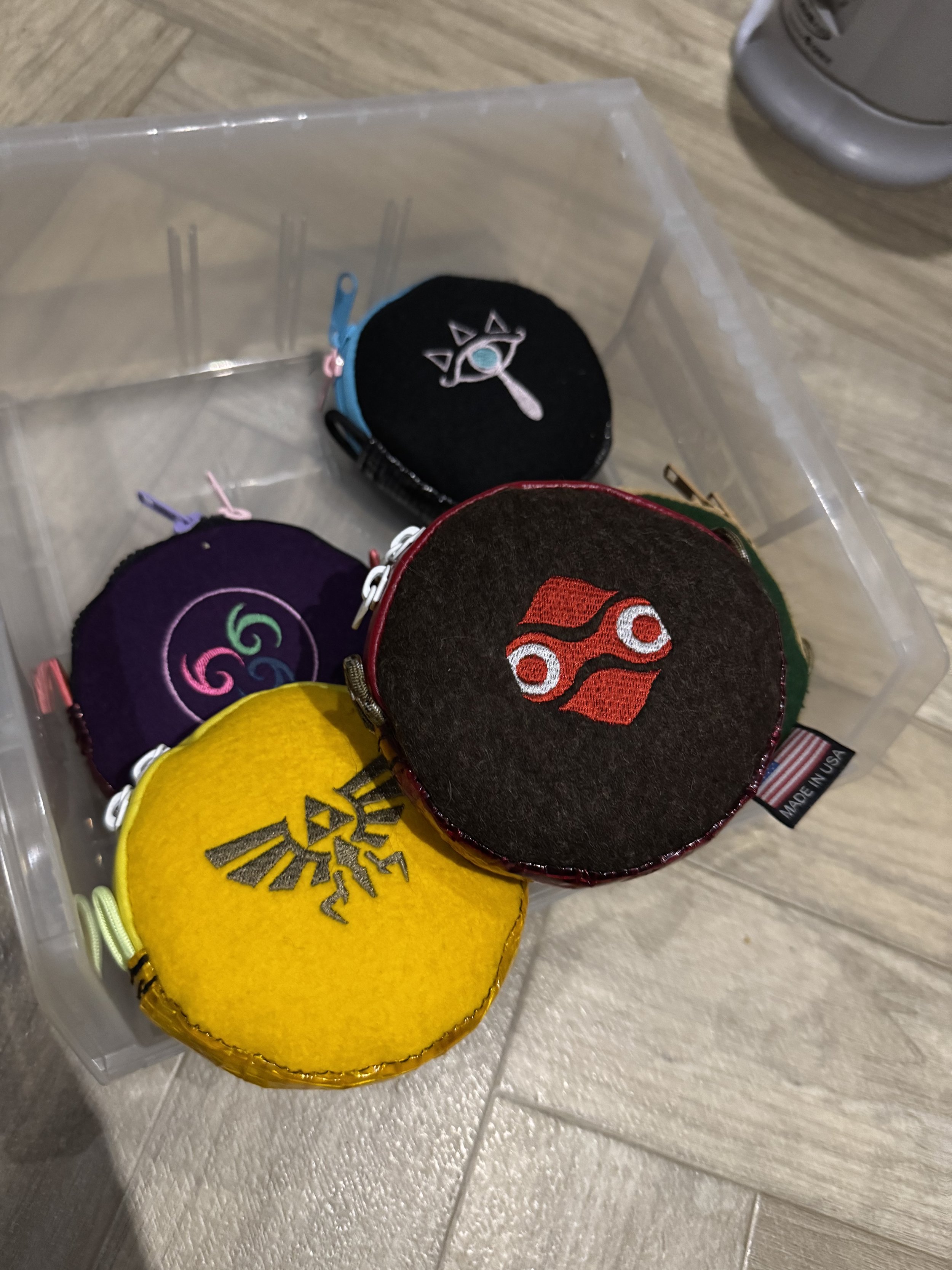 Collection of small, colorful plush pouches with embroidered symbols, inside a clear plastic storage box on a wooden floor.