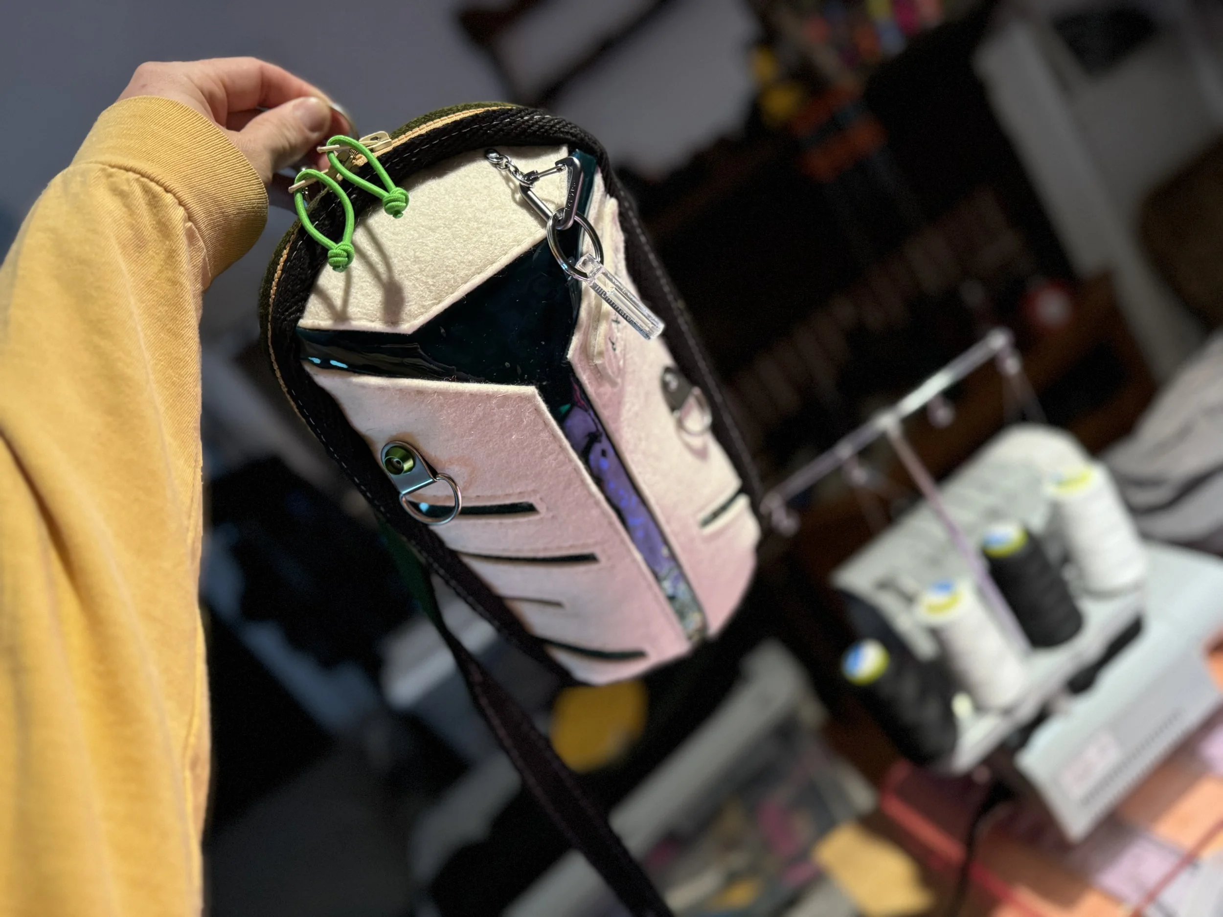 A close-up of a backpack with various hooks and clips, held by a person wearing a yellow long-sleeve shirt, in a store with shelves of thread and other sewing supplies in the background.