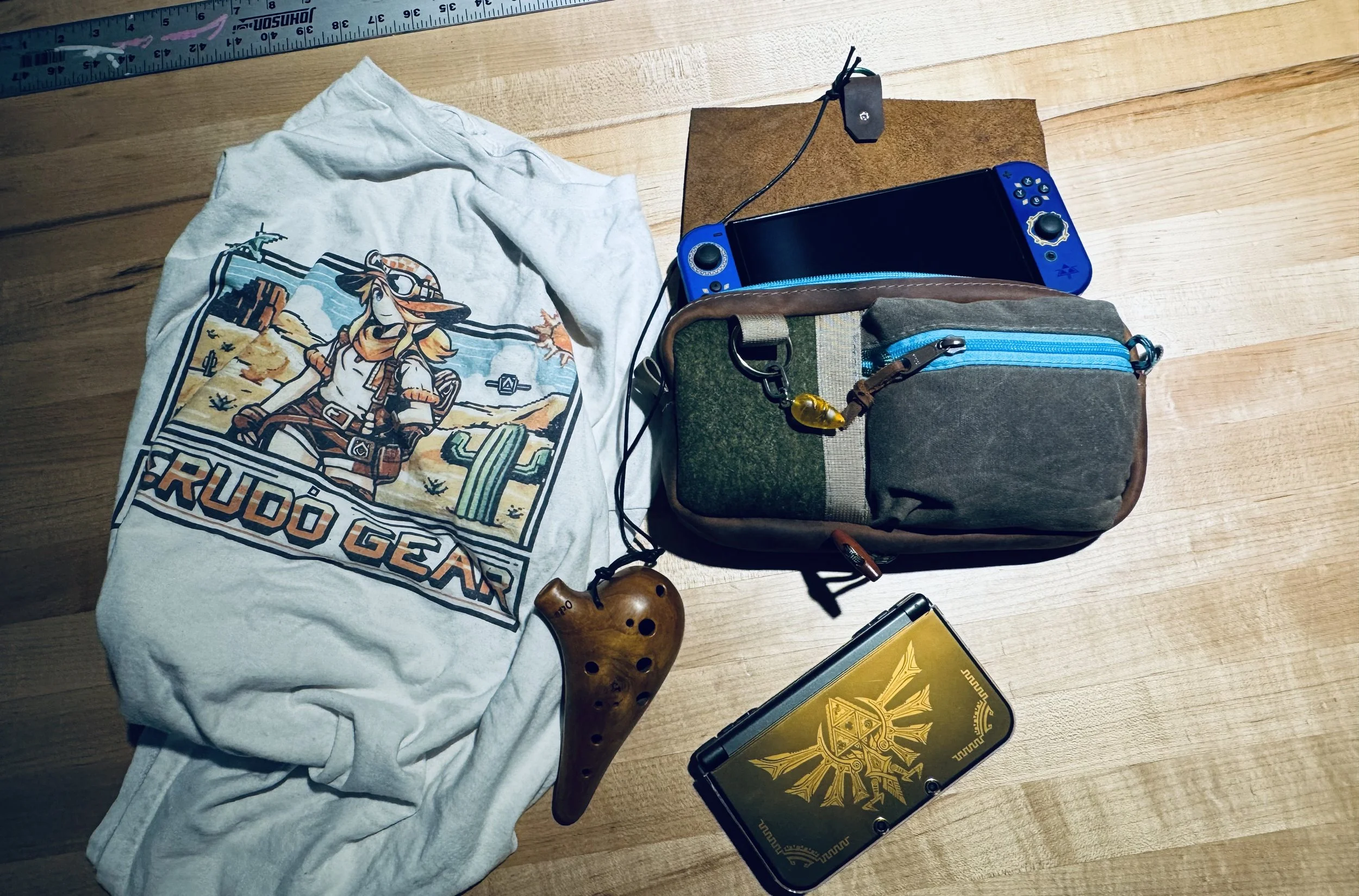 A white T-shirt with a graphic of a cowgirl in a desert landscape, a wooden ocarina, a portable gaming device with a gold design, a multi-colored bag, a blue Nintendo Switch OLED, and fabric-covered wooden surface with measurement rulers.