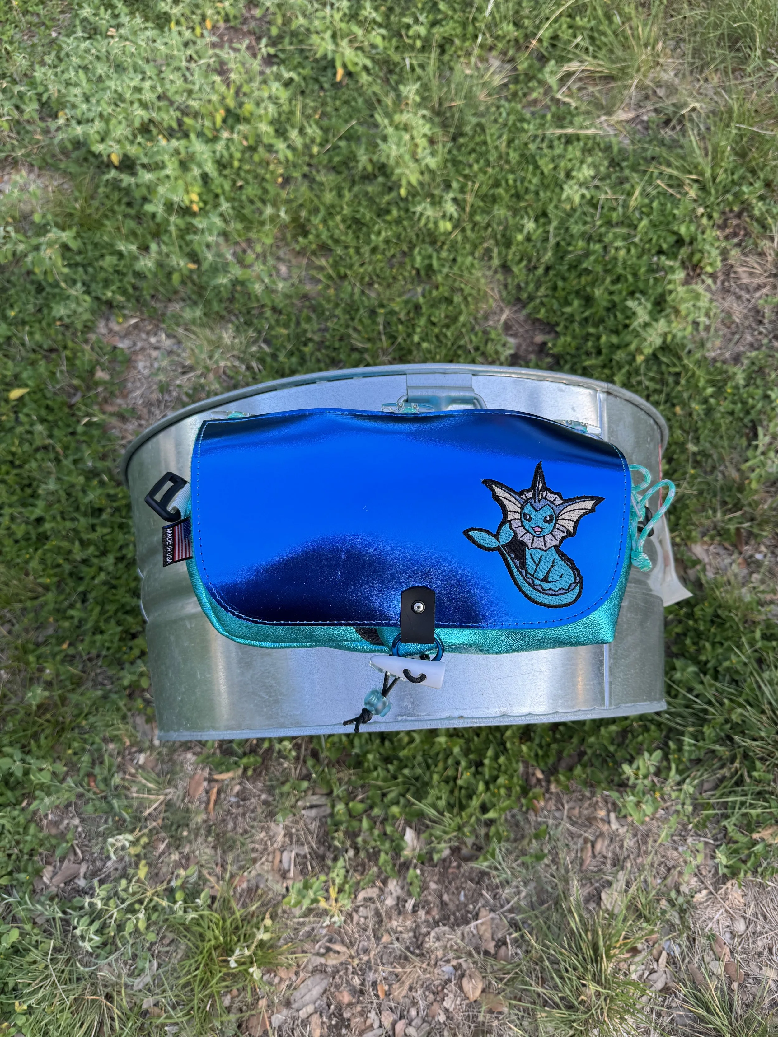 A metallic silver helmet with a blue pouch attached on top, featuring an embroidered Pokémon character, Vaporeon, in a grassy outdoor area.