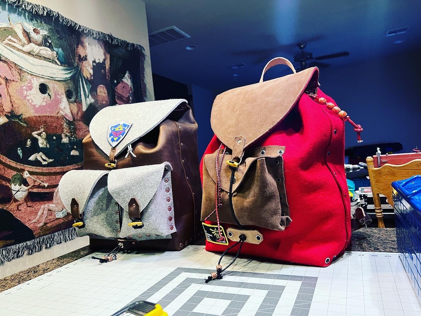 Three backpacks made of leather and fabric with multiple pockets and closures, displayed on a table with a tiled surface, with a colorful tapestry and a dark room in the background.