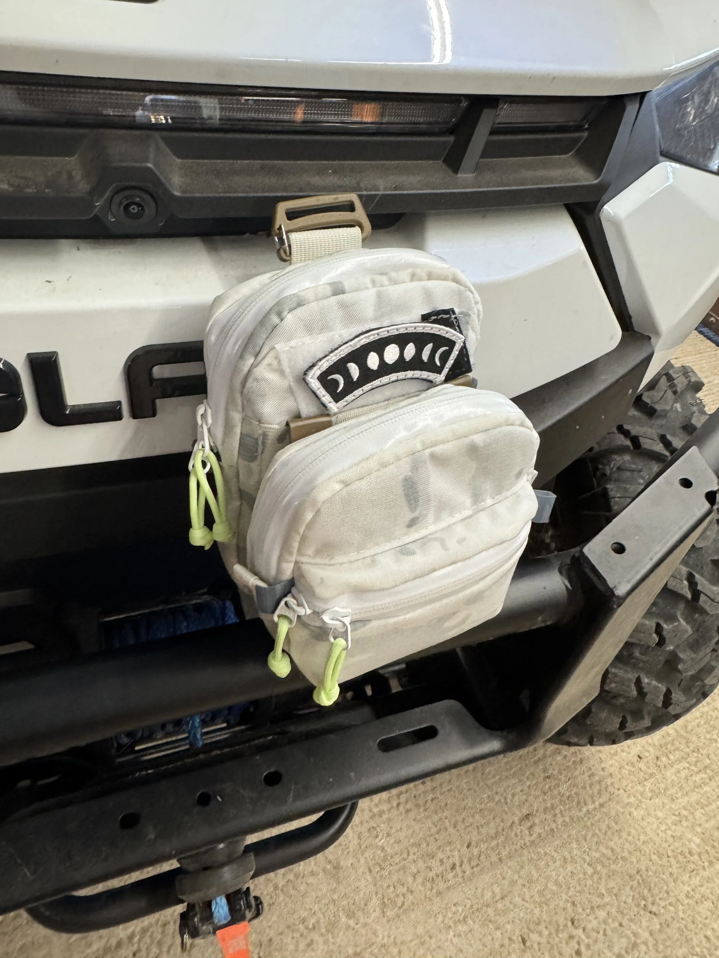 A small, beige backpack hanging on the front of a vehicle, with a black patch featuring a semi-circular design. The backpack has two zippers with green zipper pulls.