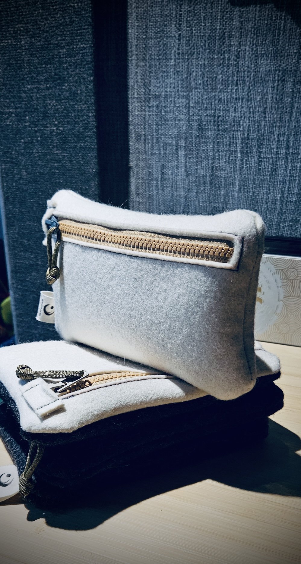 Three small felt pouches, one white with a gold zipper, one gray with a gold zipper, and black, stacked on a light wooden surface with a dark fabric background.