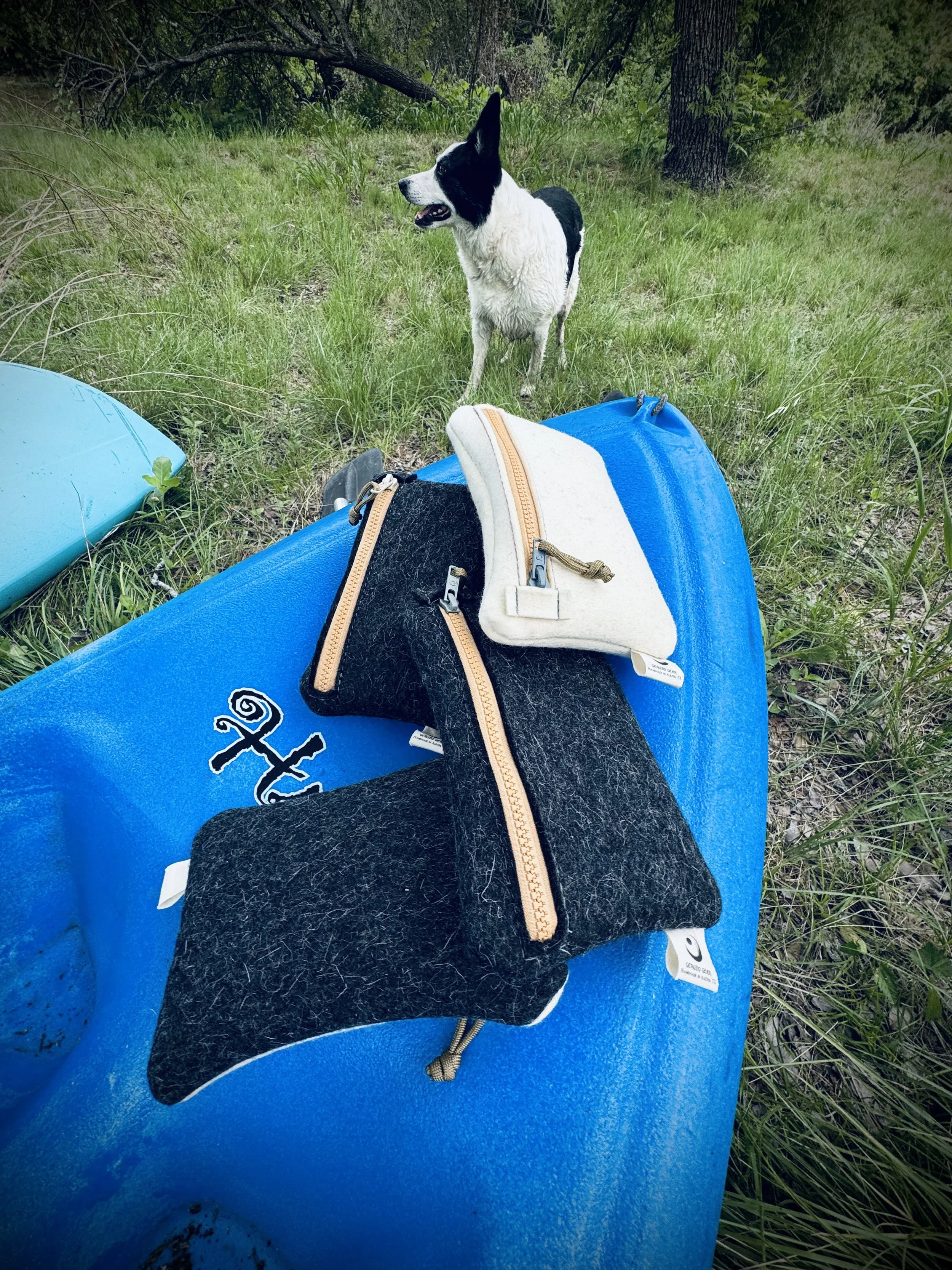 A black and white dog standing in grass near a blue kayak with three small zippered pouches in black and white placed on top.