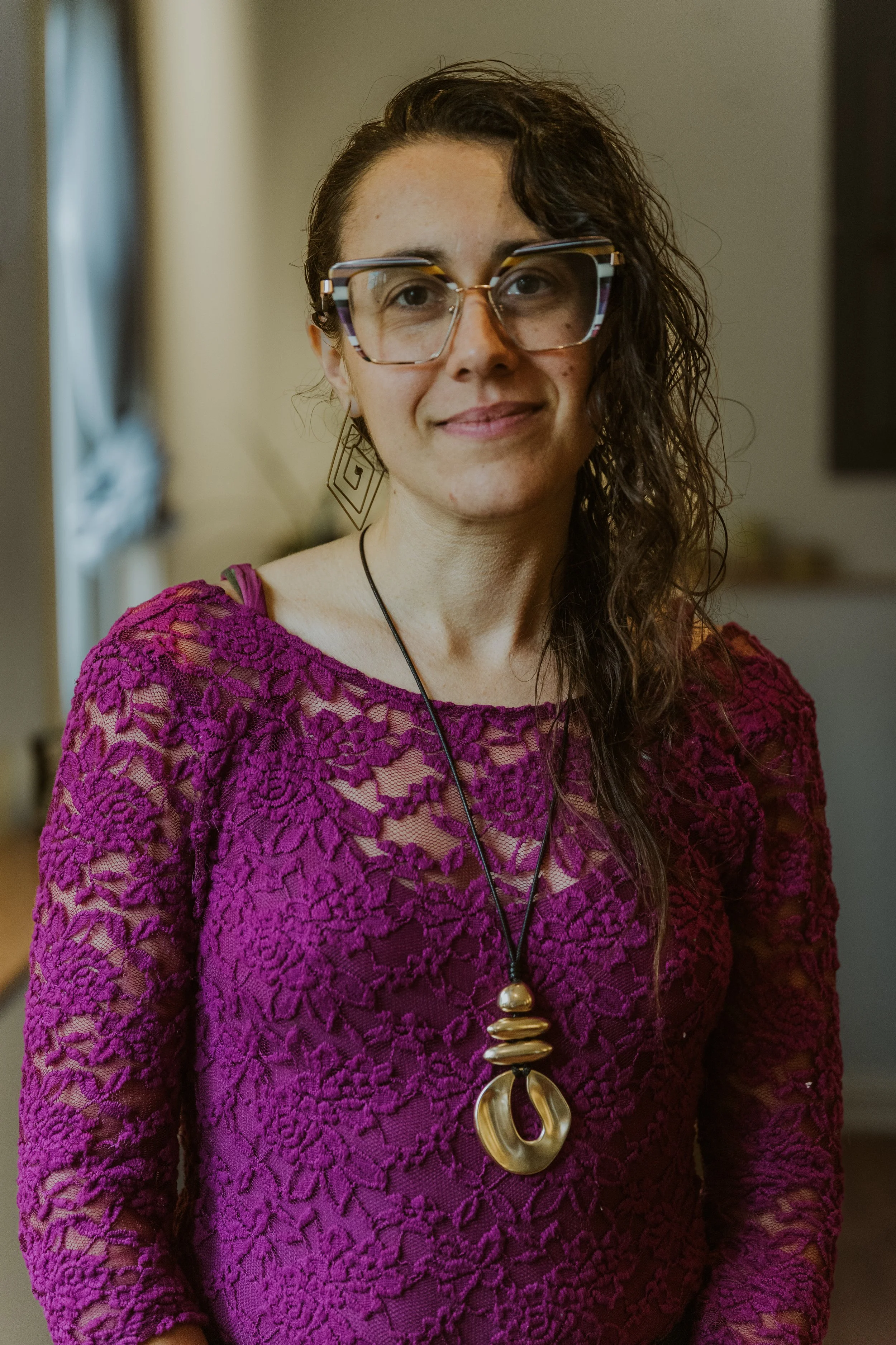 Caspian, a person with curly hair wearing large glasses, gold earrings, and a purple lace top with a large statement necklace in Northeast Minneapolis at Minnesota Integrative Health Studio.