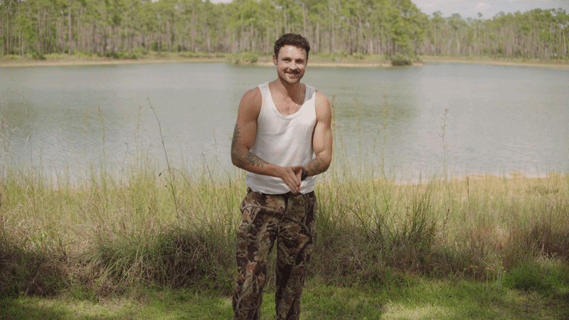 A smiling man with tattoos on his arms standing outdoors near a body of water and grass, with trees in the background.