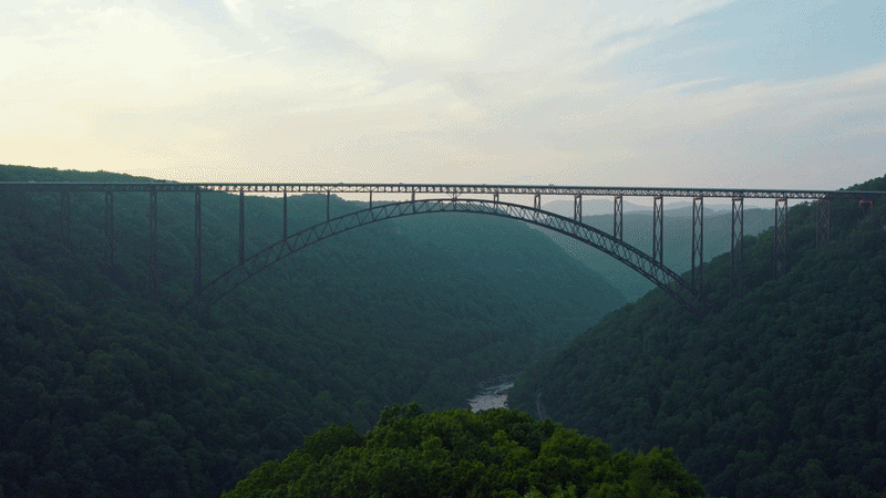 Steel arch bridge spanning a verdant valley with a river below, under a cloudy sky.