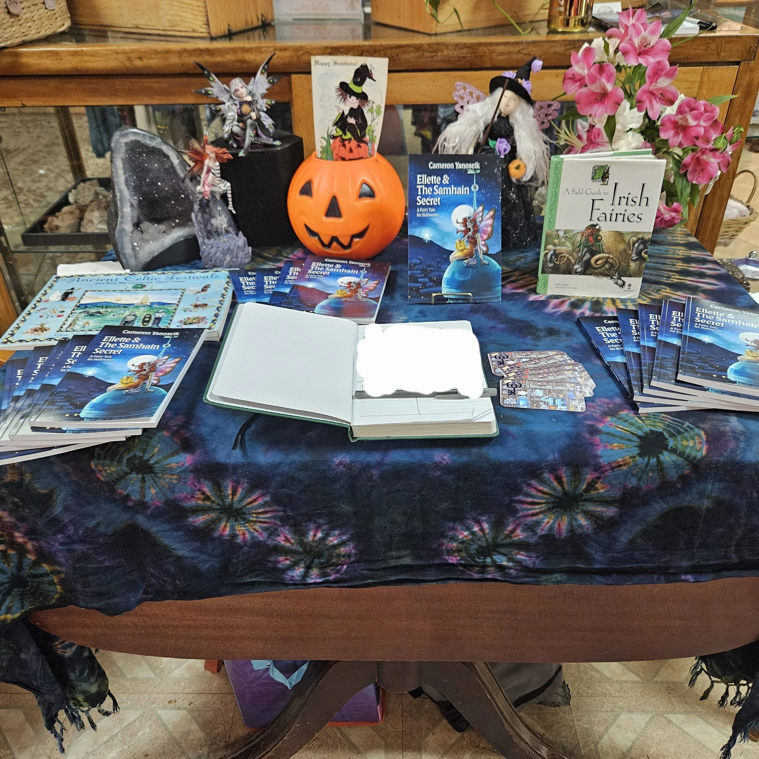 A table decorated with Halloween and Irish fairy themed items, including fairy dolls, a pumpkin container with a Halloween card, a book titled 'A Field Guide to Irish Fairies,' brochures of 'Ellette & The Samhain Secret,' brochures, a notebook, a flower arrangement, and a colorful cloth covering the table.