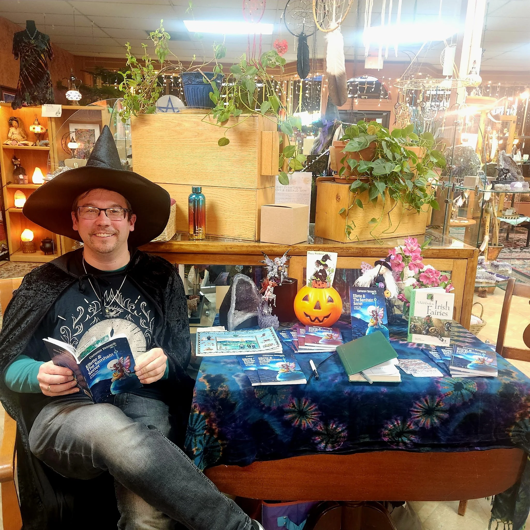 The author dressed as a wizard with a large black hat, black cape, and glasses, sitting at a table with books, a pumpkin-shaped lantern, and decorative objects in a store decorated for Halloween during one of his book reading events.
