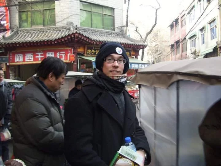 A young man wearing glasses, a black jacket, and a black beanie with a Pittsburgh Steelers logo, standing outdoors among other people, in front of a street scene with buildings and a traditional Chinese-style structure.