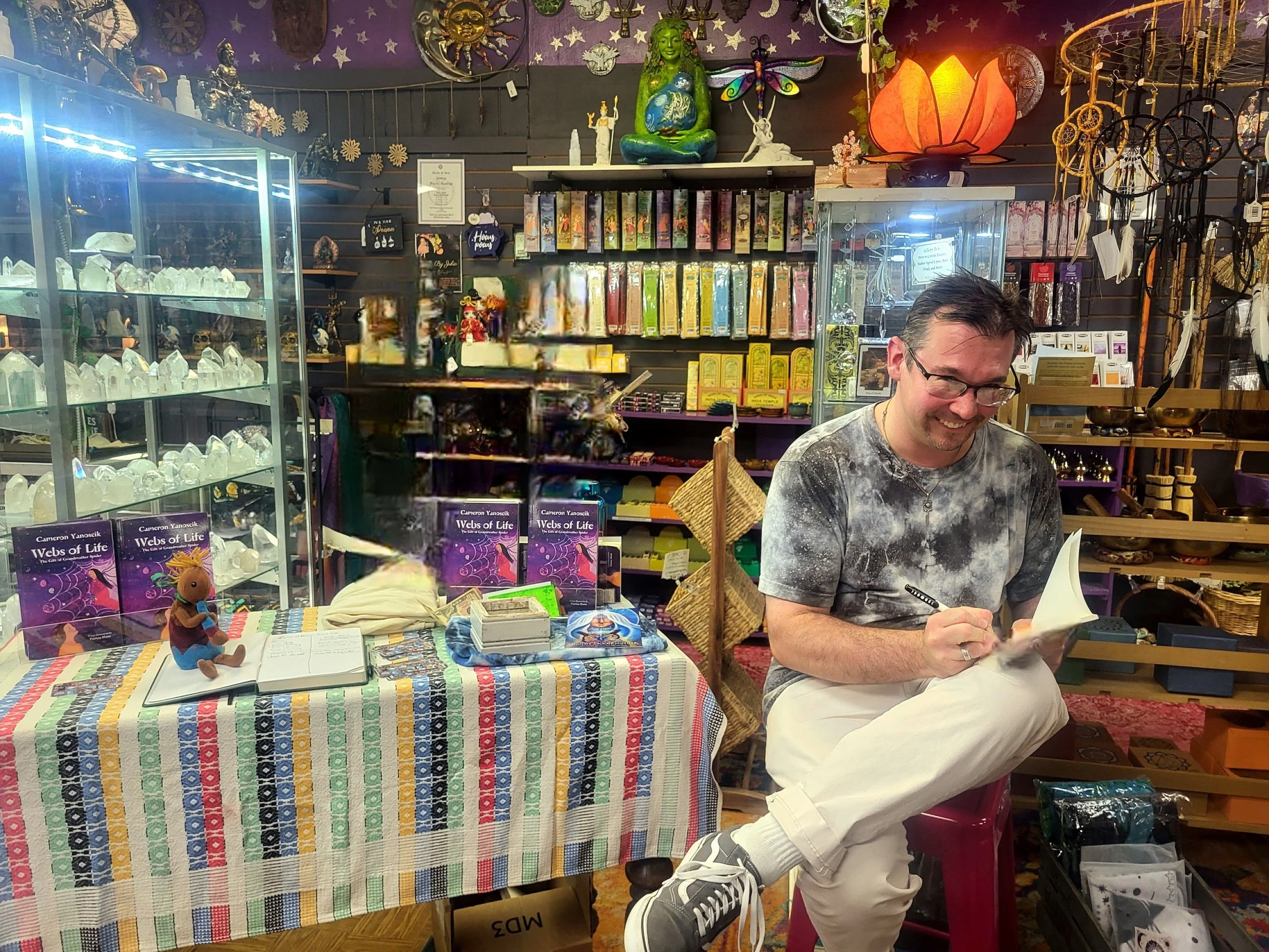 A man with glasses, smiling, wearing a tie-dye shirt and white pants, sitting on a pink stool in a store, signing a book. The store has shelves with crystals, decorative items, and books, with a colorful display table in front.
