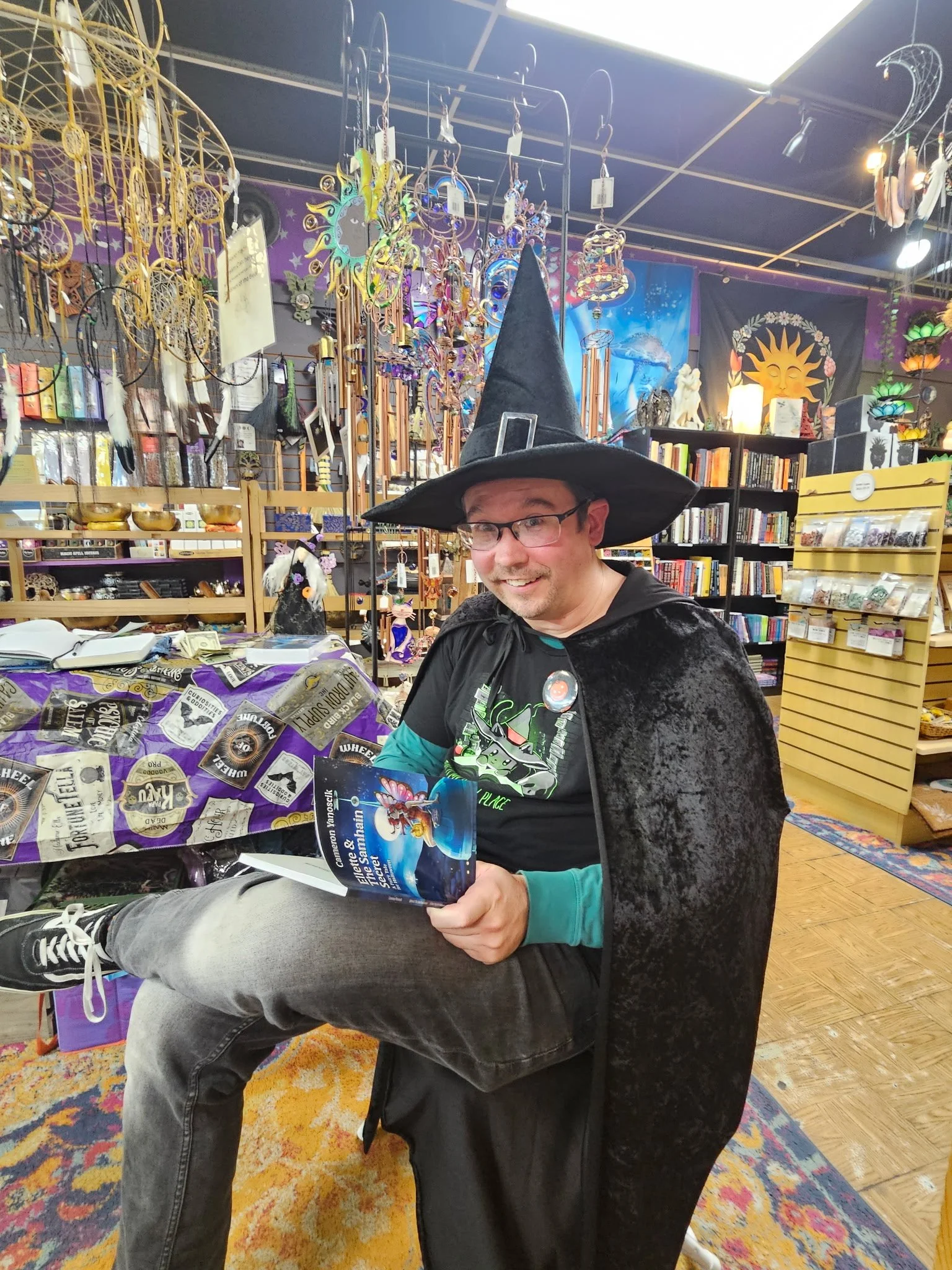 The author as a wizard, wearing a tall black hat and a black cape, sitting in a witchy gift shop, holding a his newly published Halloween book and smiling at the camera.