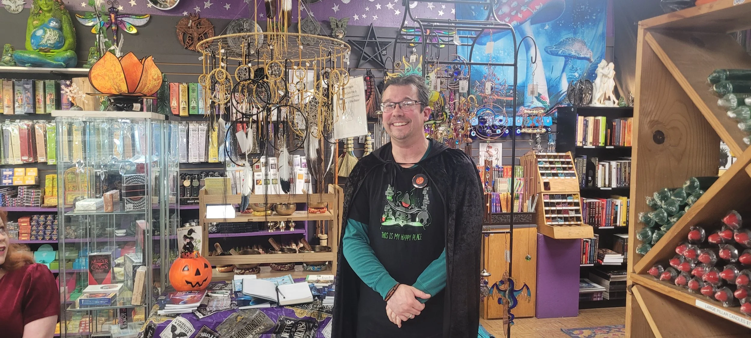 The author standing inside a store filled with colorful merchandise, including jewelry, books, Halloween decorations, and candles, after reading of his Halloween fairy tale book, newly published.