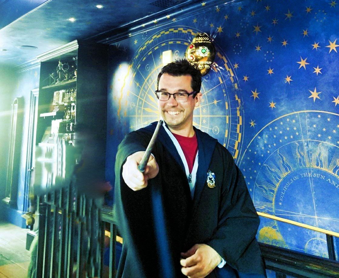 Man dressed as Harry Potter holding a wand in a themed room with a blue cosmic wall and star patterns.