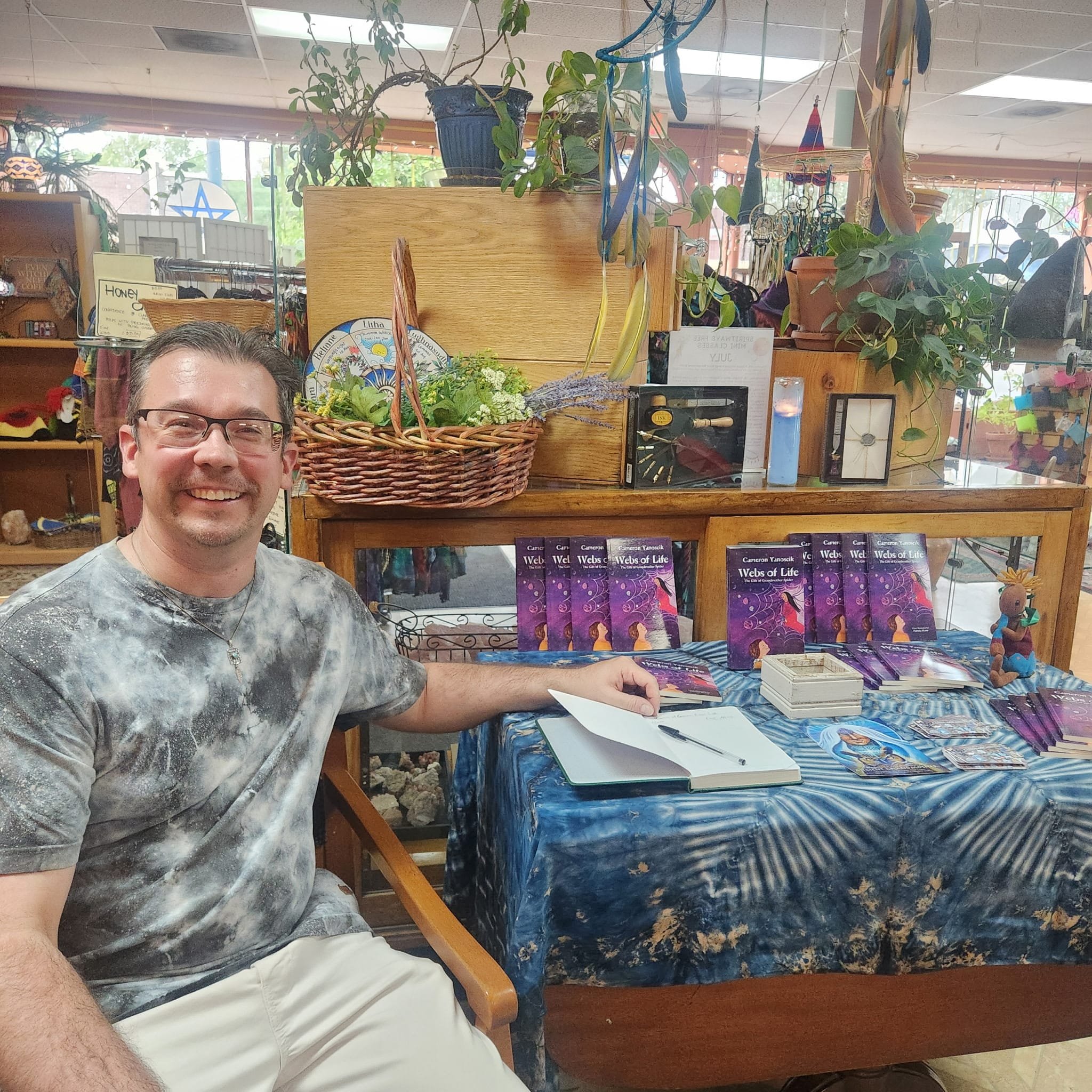 A man sitting at a table with books and tarot cards, smiling for the camera inside a store with plants and decorative items.