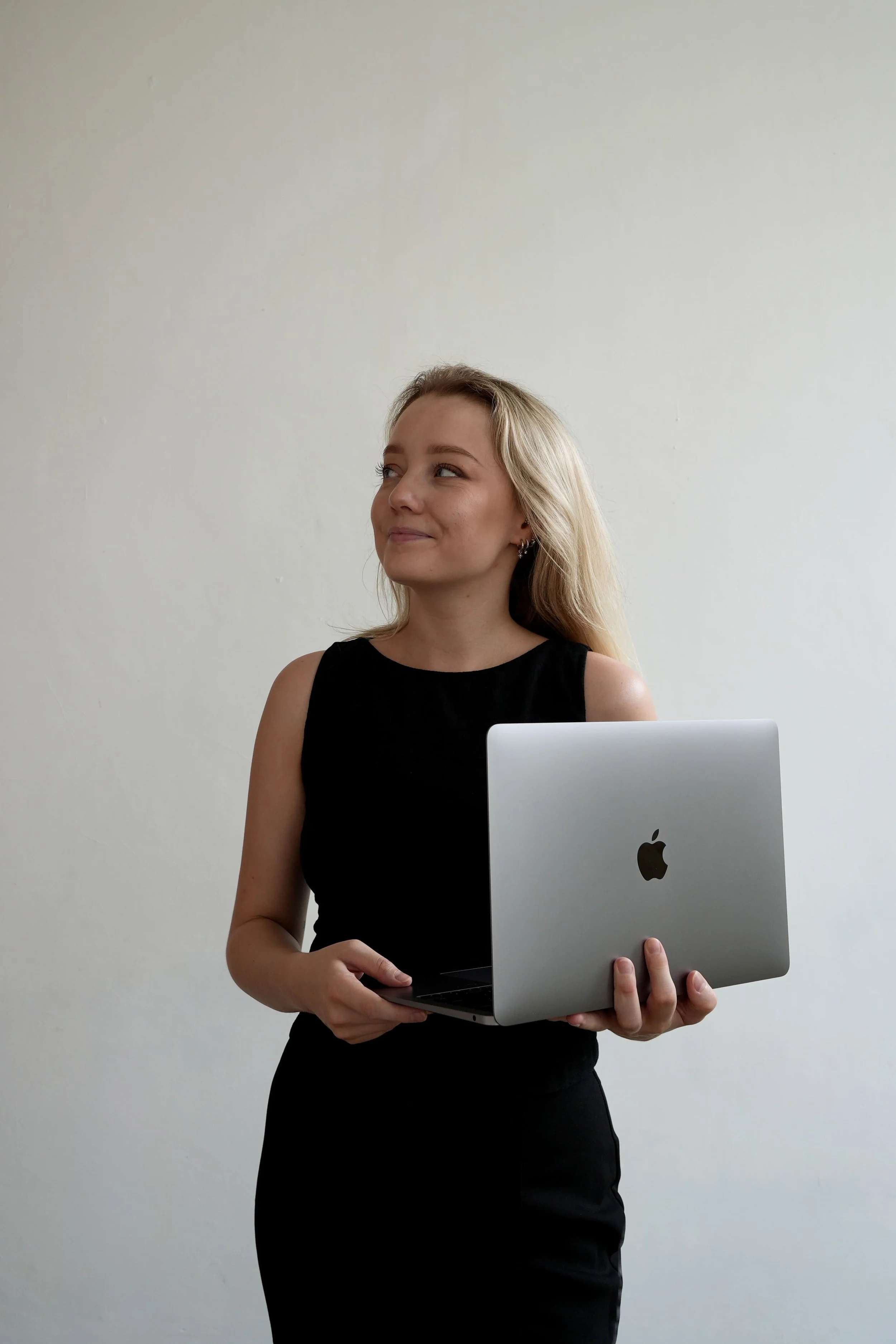 Rachel is wearing a black sleeveless top holding a silver MacBook laptop in front of a plain white wall.