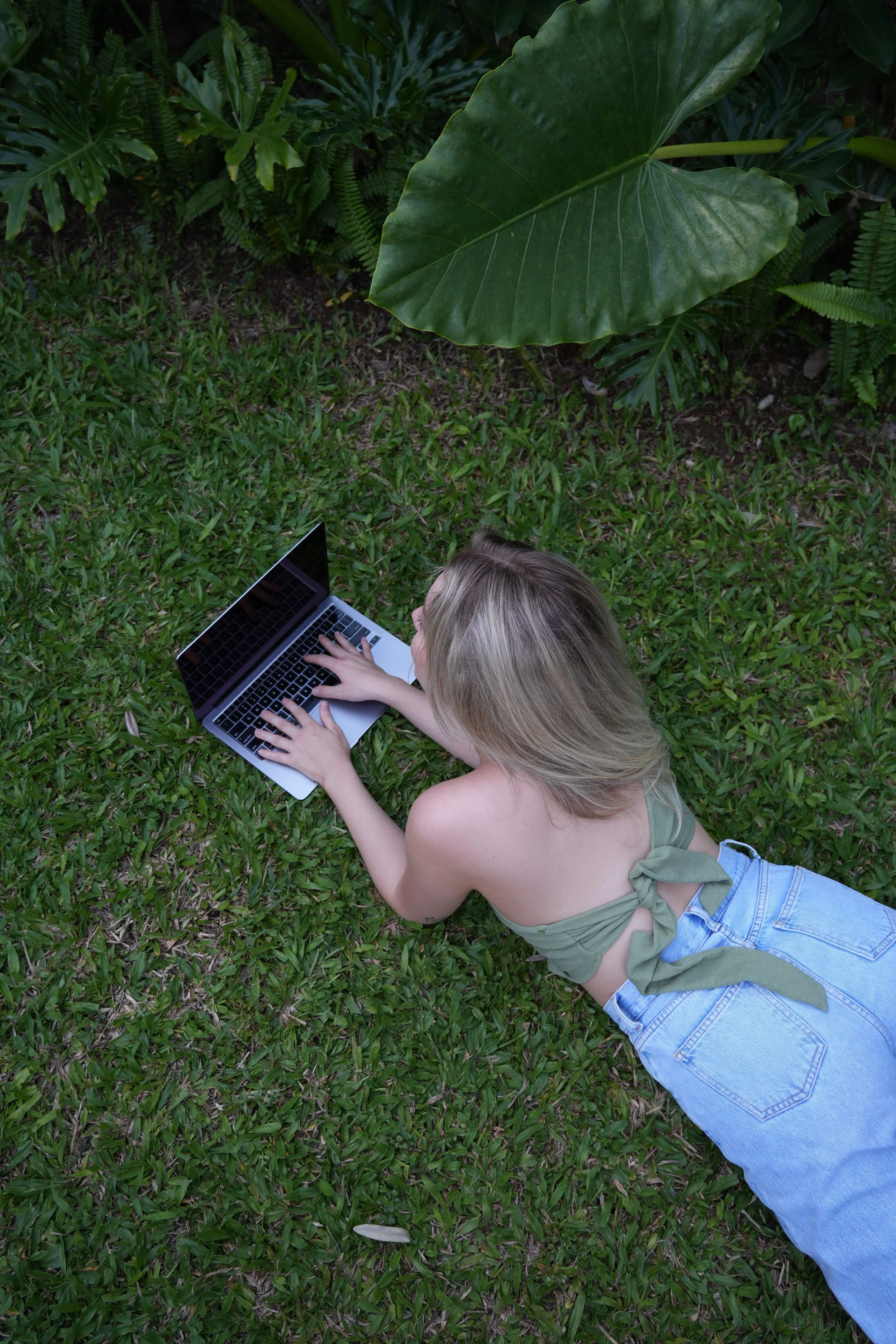 Rachel is lying on green grass, typing on a laptop, surrounded by large tropical leaves.