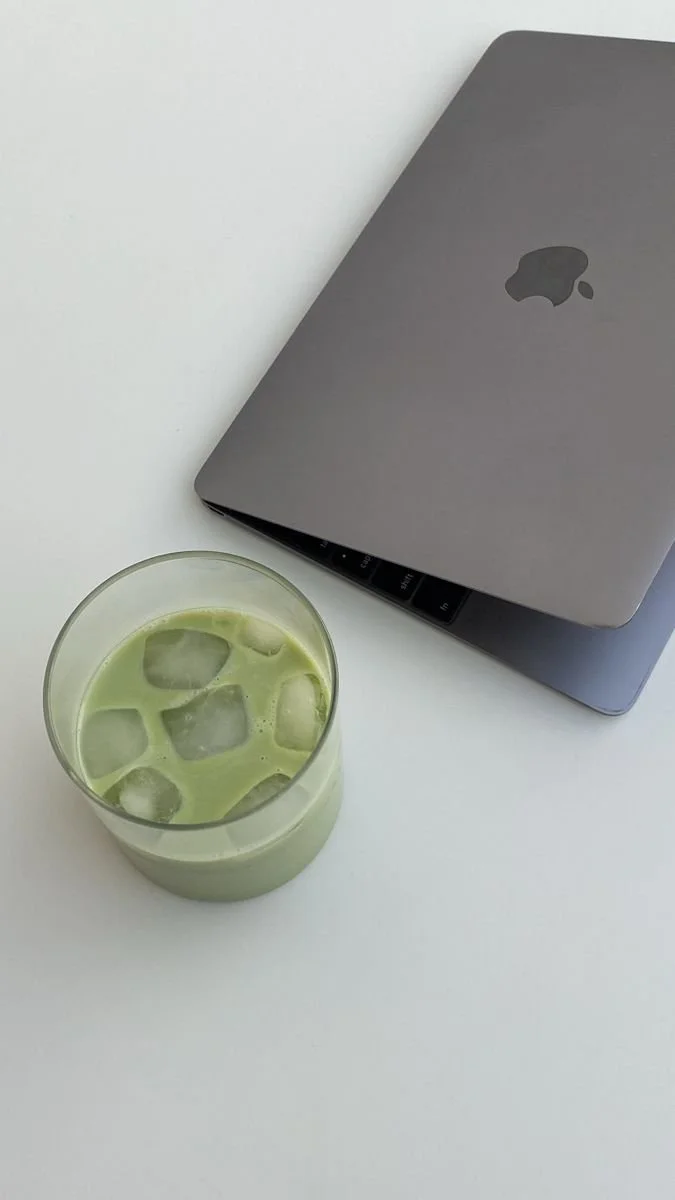 A closed gray MacBook with an Apple logo and a glass of green iced matcha latte with ice cubes on a white surface.