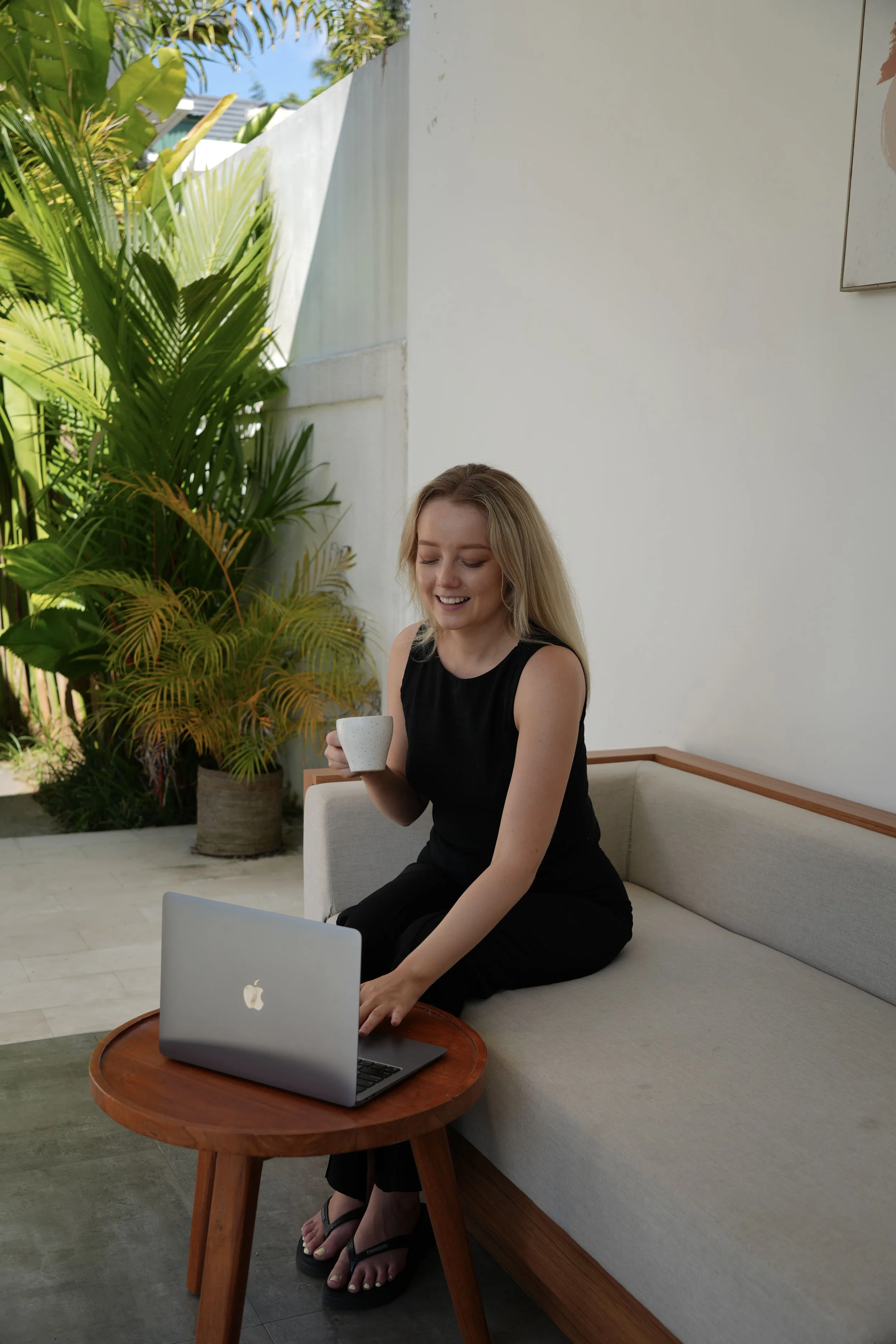 Rachel sits on a beige outdoor couch, holding a mug and looking at her laptop open on a small round wooden table in front of her.