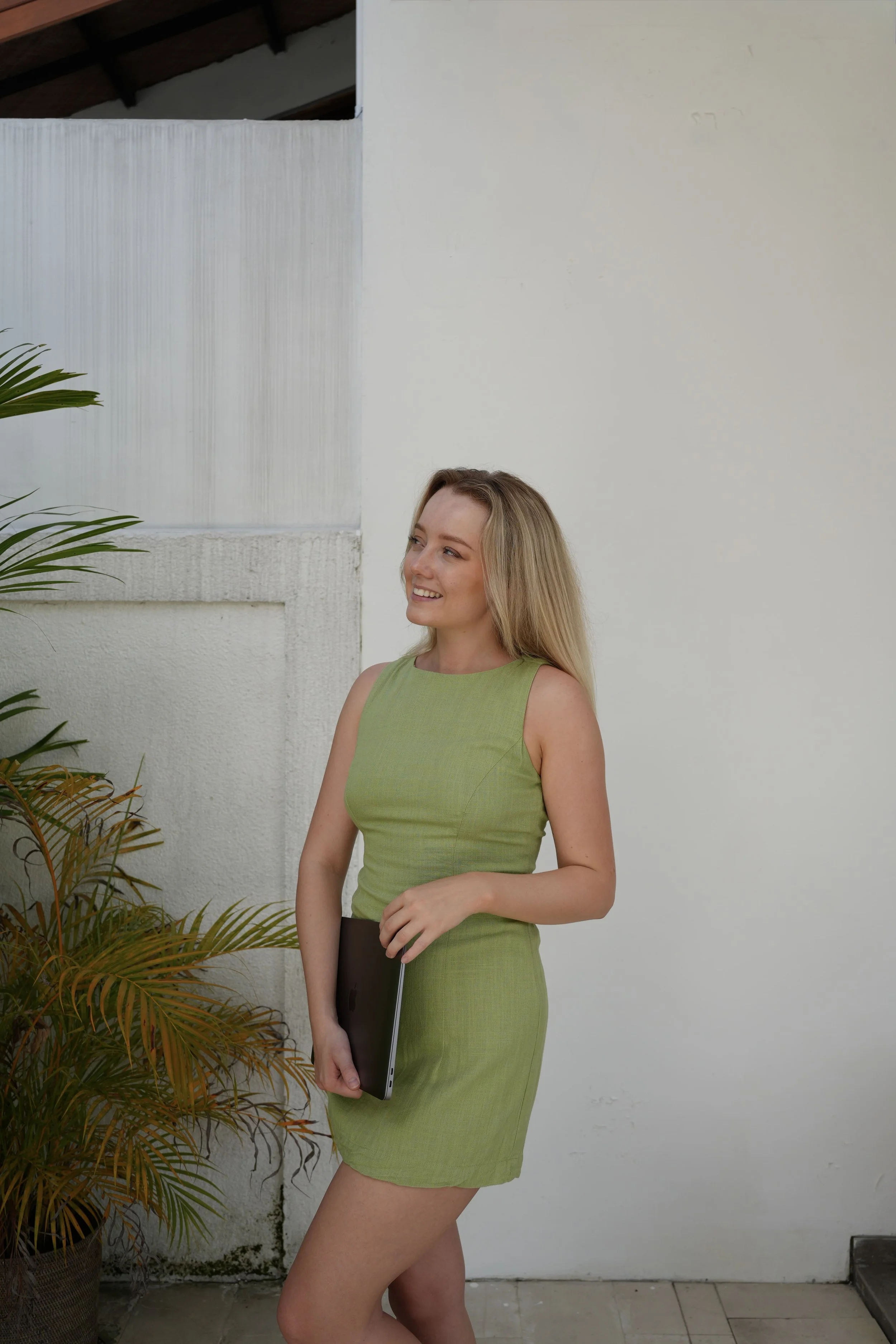 Rachel is wearing a green sleeveless dress, holding a closed laptop, standing outdoors near a white wall with plants nearby, smiling and looking to her left.