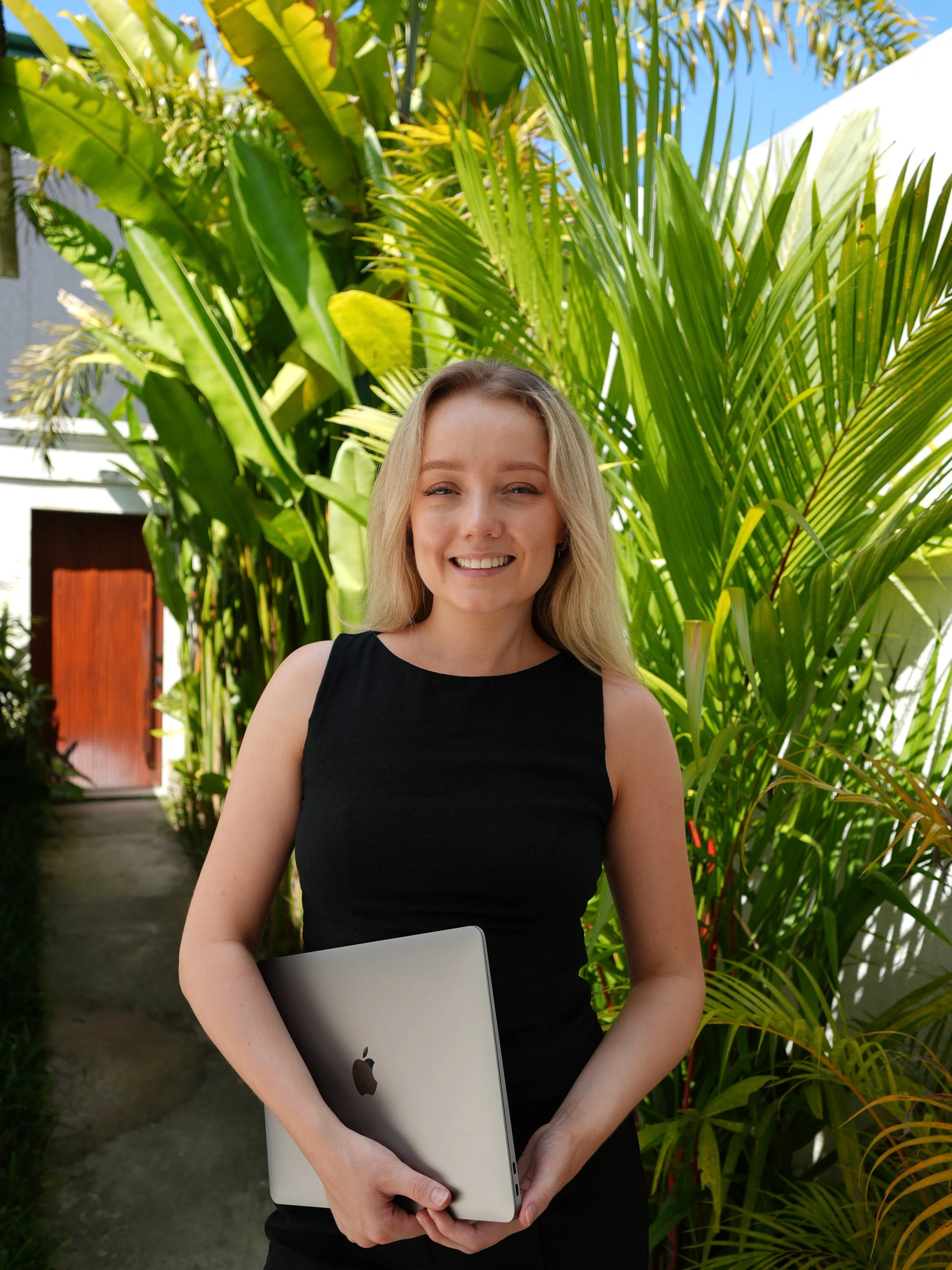 Rachel is standing outdoors in front of lush green tropical plants, holding a closed silver laptop with an Apple logo, smiling at the camera.