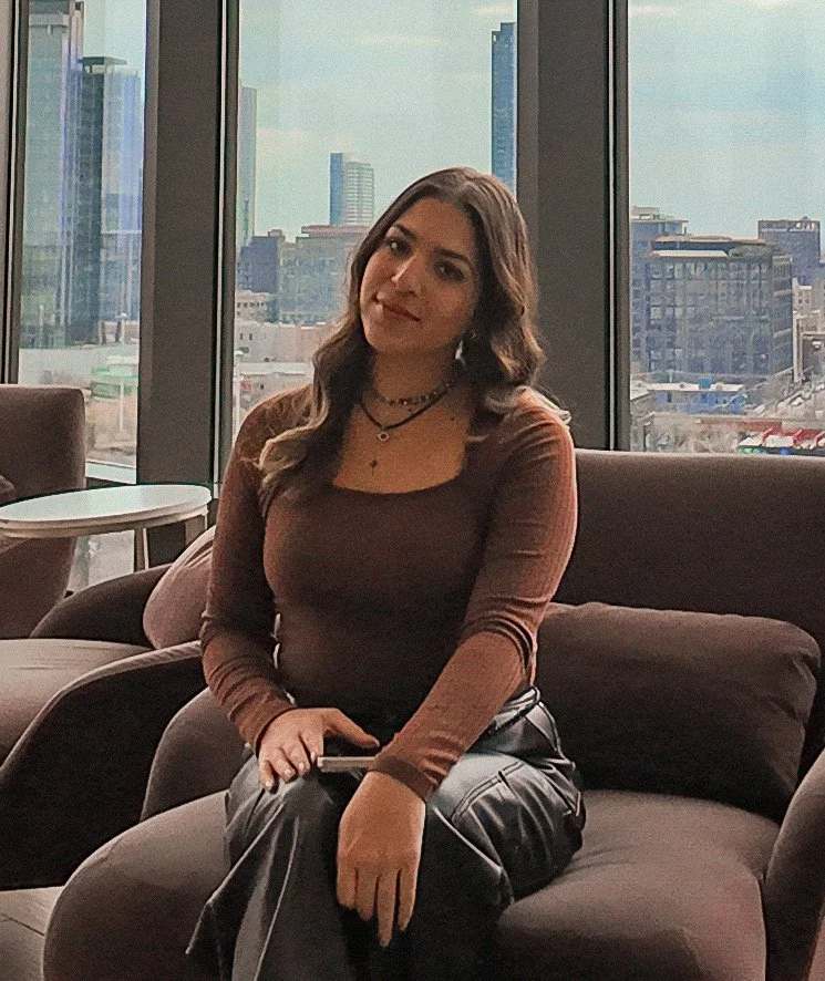 Jazmin, Rachel's client, sitting on a dark sofa in a modern high-rise building with large windows and a city skyline view, holding a phone.