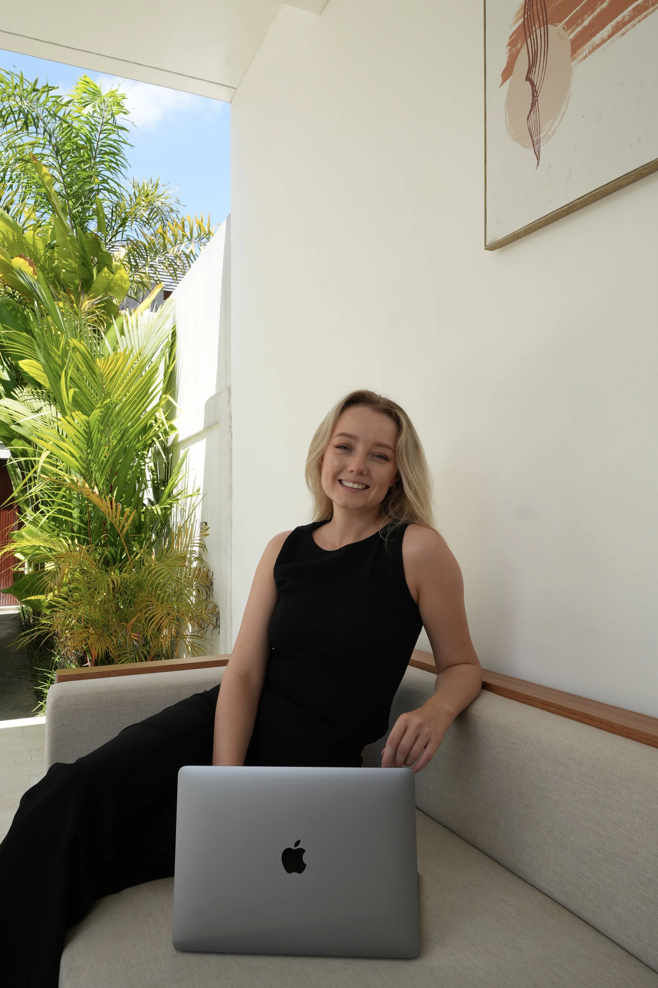 Rachel is sitting on a beige couch with a laptop in front of her, smiling, in a bright room with large green plants outside the window.