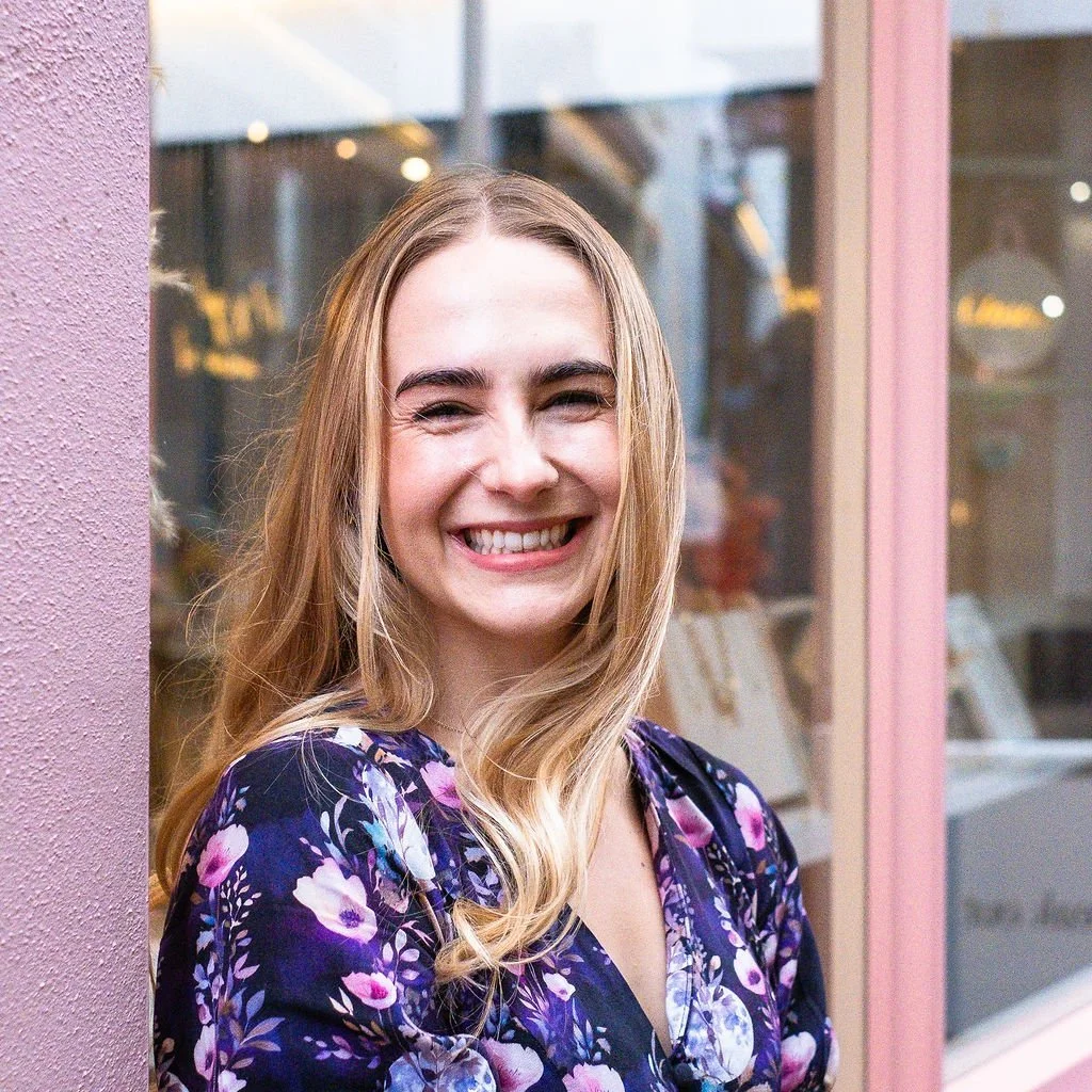 Ella, Rachel's client, is smiling, she has long, wavy blonde hair and is standing outside near a pink wall and glass window.